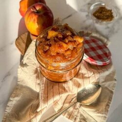 apples caramelized in a jar, over a striped towel, a spoon, apples and brown sugar on the sides, over a marble background