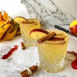 apple fizz mocktail with apple slices and cinnamon sticks on top, apple peels, cinnamon sticks, knife, towel and leaves around, on marble background, glass bottle of ginger beer