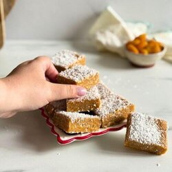 hand holding a slice of apricot slice, on a plate more apricot slice, behind a bowl of apricots, towel and two wooden boards on a marble background