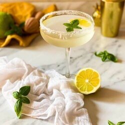 basil lemon martini in a glass on a marble background, on the side a lemon, some basil leaves and a white towel, in the background a yellow towel, lemon squeezer and cocktail shaker