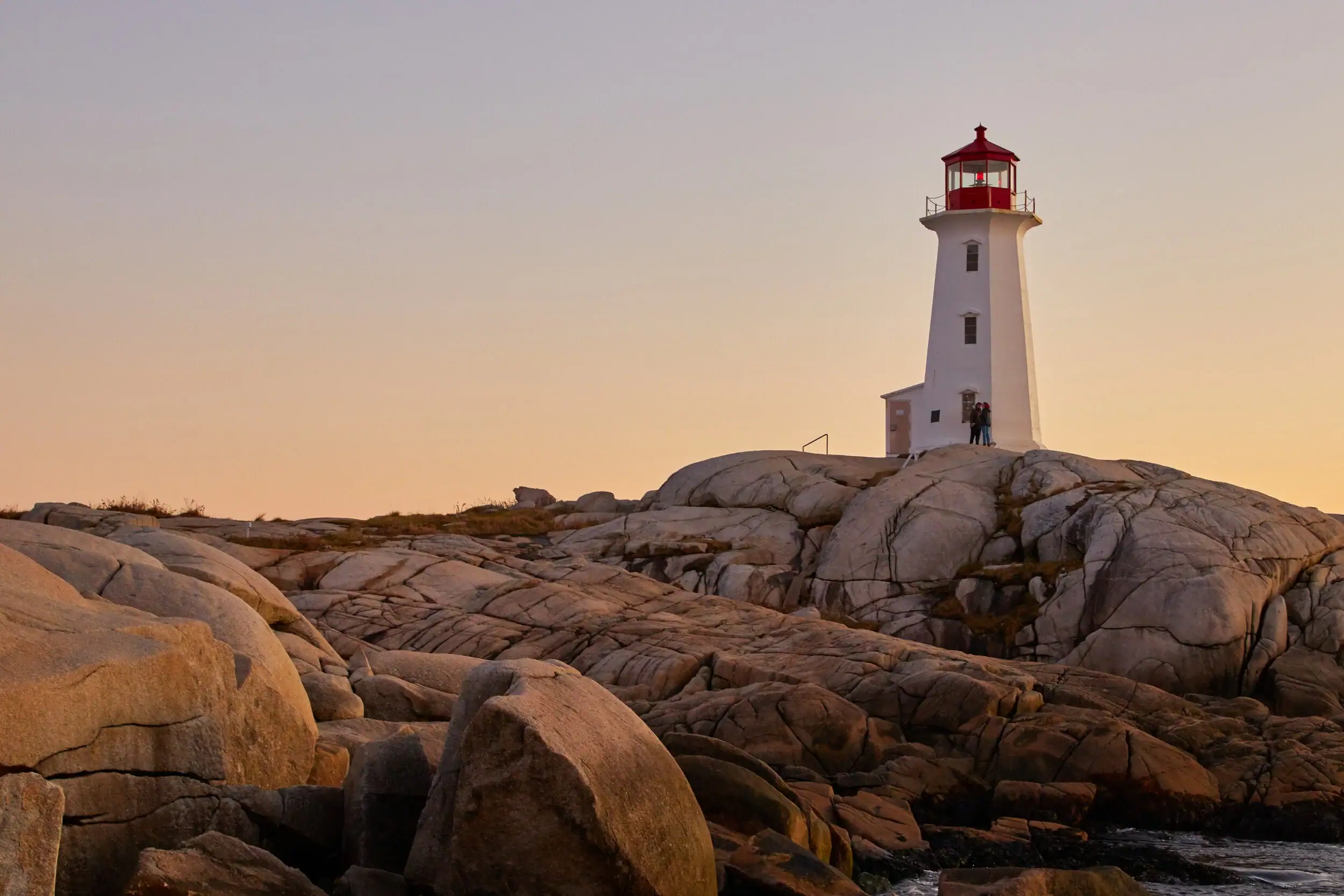 White lighthouse with a red lantern room on a rocky coastline during sunset, with two people near the base.