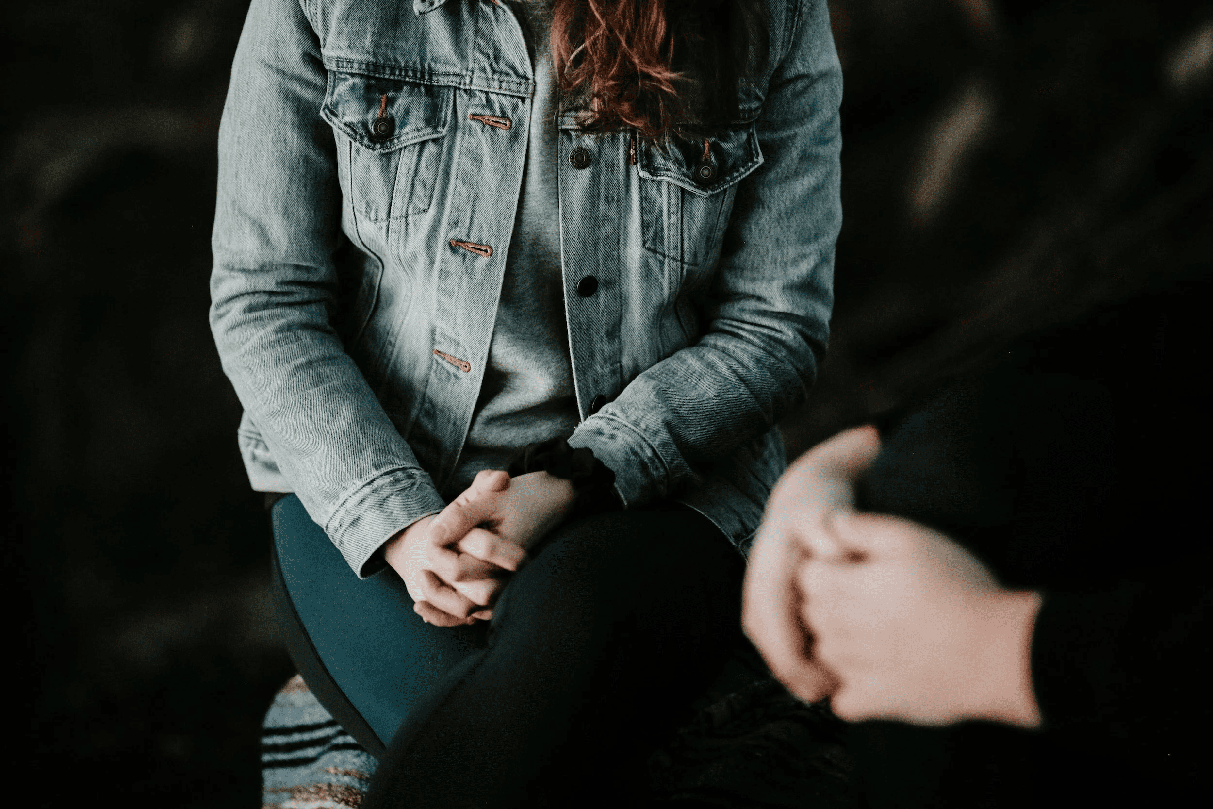 Person wearing a light denim jacket sits with hands folded in their lap; another person is blurred in the foreground representing doing online therapy in Michigan