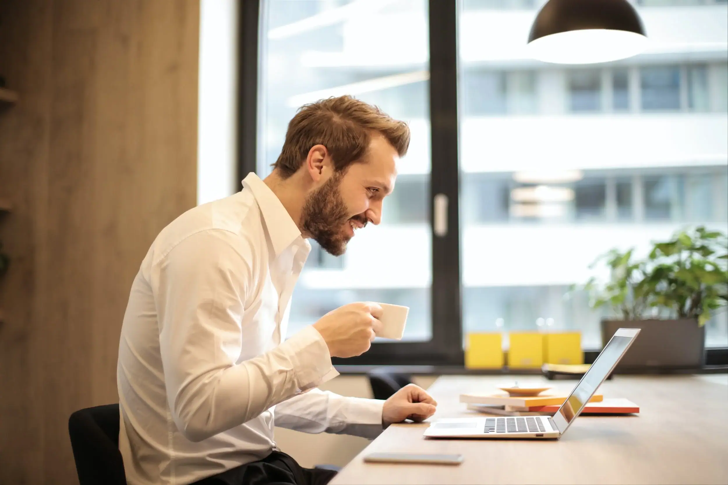 Man in a white shirt sits at a desk in an office, holding a white mug while looking at a laptop screen. Start online therapy michigan today.