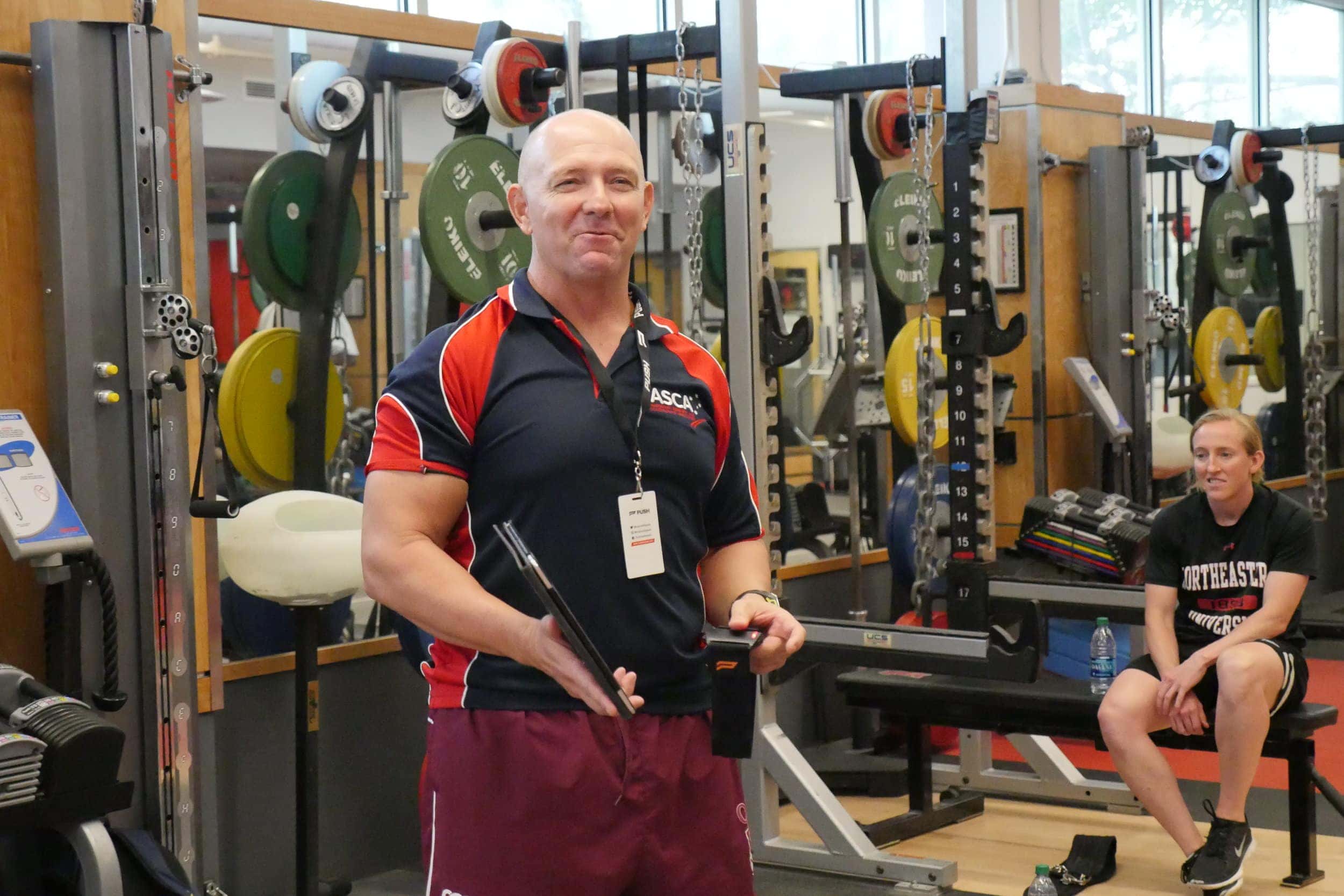A strength and conditioning coach standing confidently in a gym, clipboard in hand.