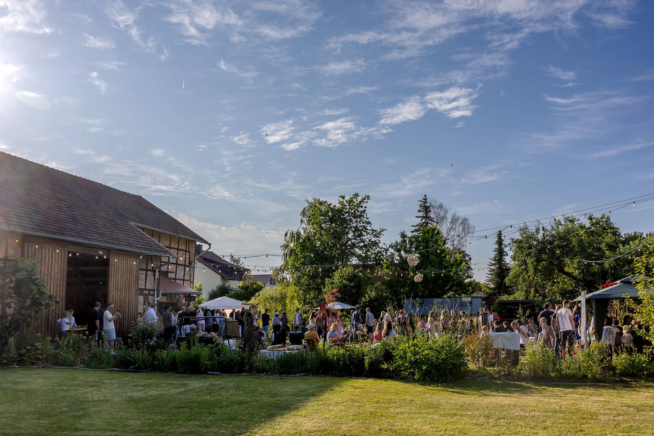 die lebhafte und offene Atmosphäre des Kultursommers, bei dem zahlreiche Gäste im grünen Garten vor der rustikalen Scheune zusammenkommen fotografiert mit der Canon EOS R5M2 & dem RF 28-70mm f2.0
