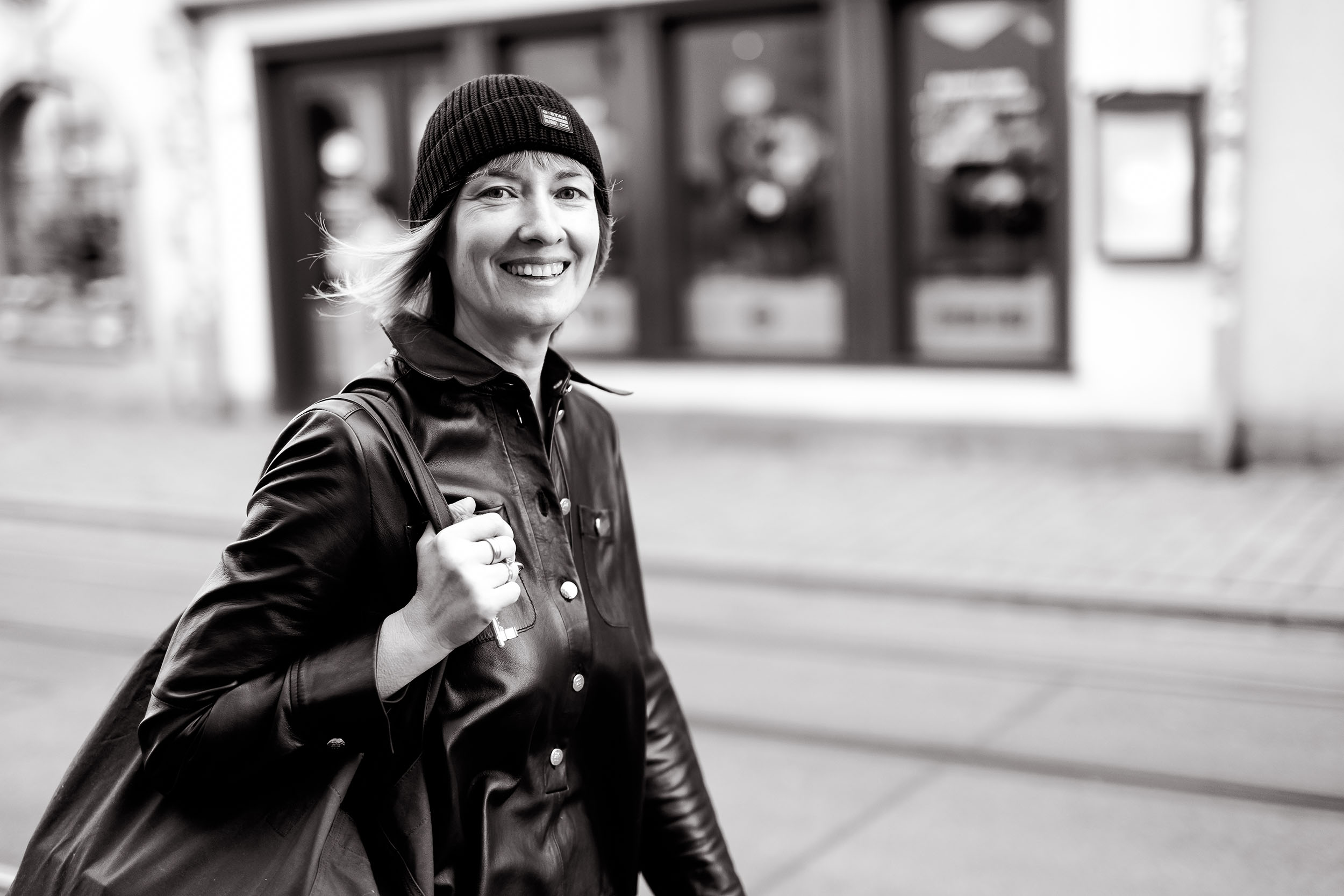 Lächelnde Frau in schwarzer Lederjacke und Mütze auf einer Einkaufstraße in seitlicher Bewegung fotografiert