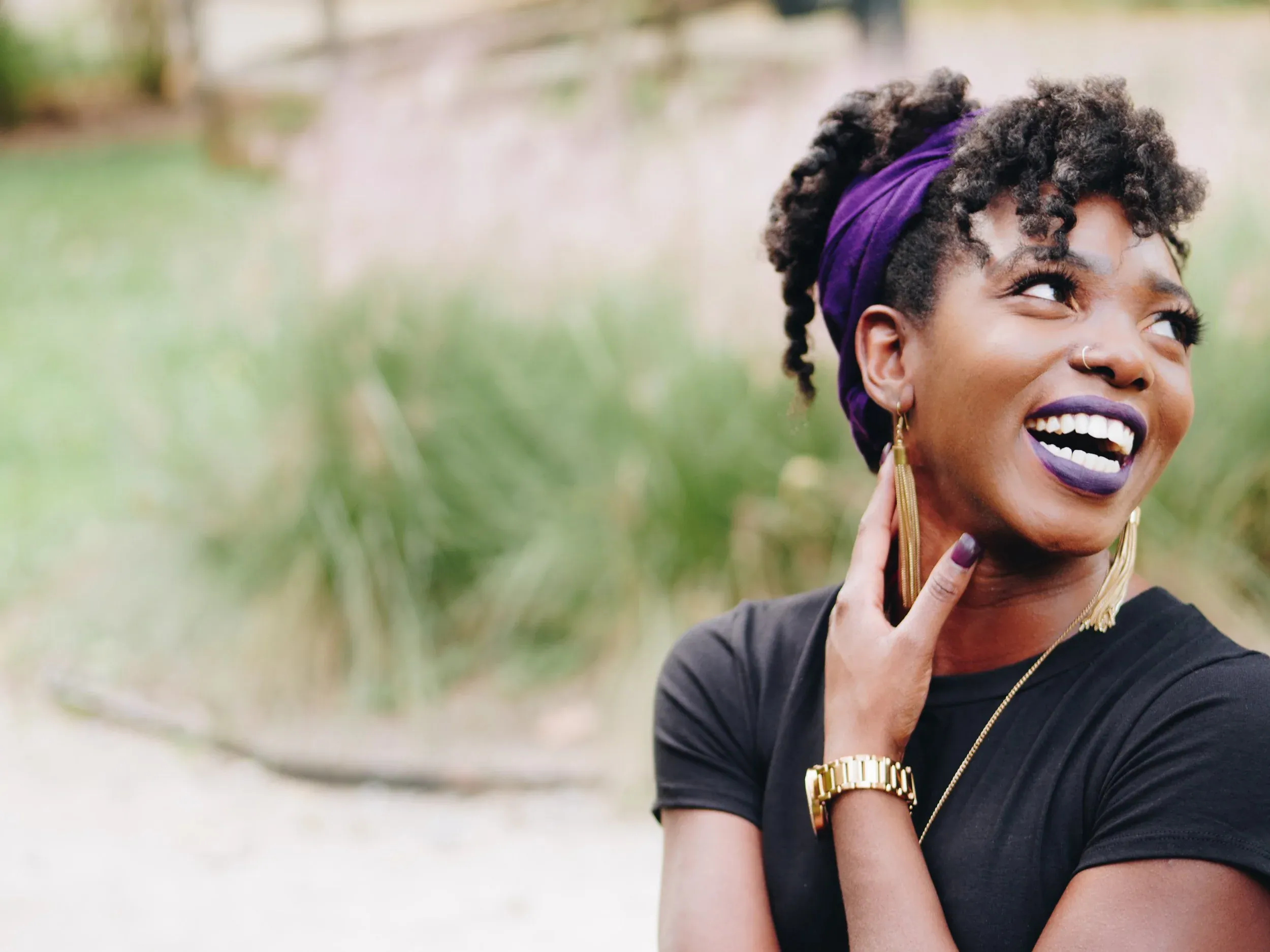 Woman smiling outdoors, wearing a purple headwrap and gold jewelry with a black top, bright expression. Start ptsd therapy in detroit, mi today!