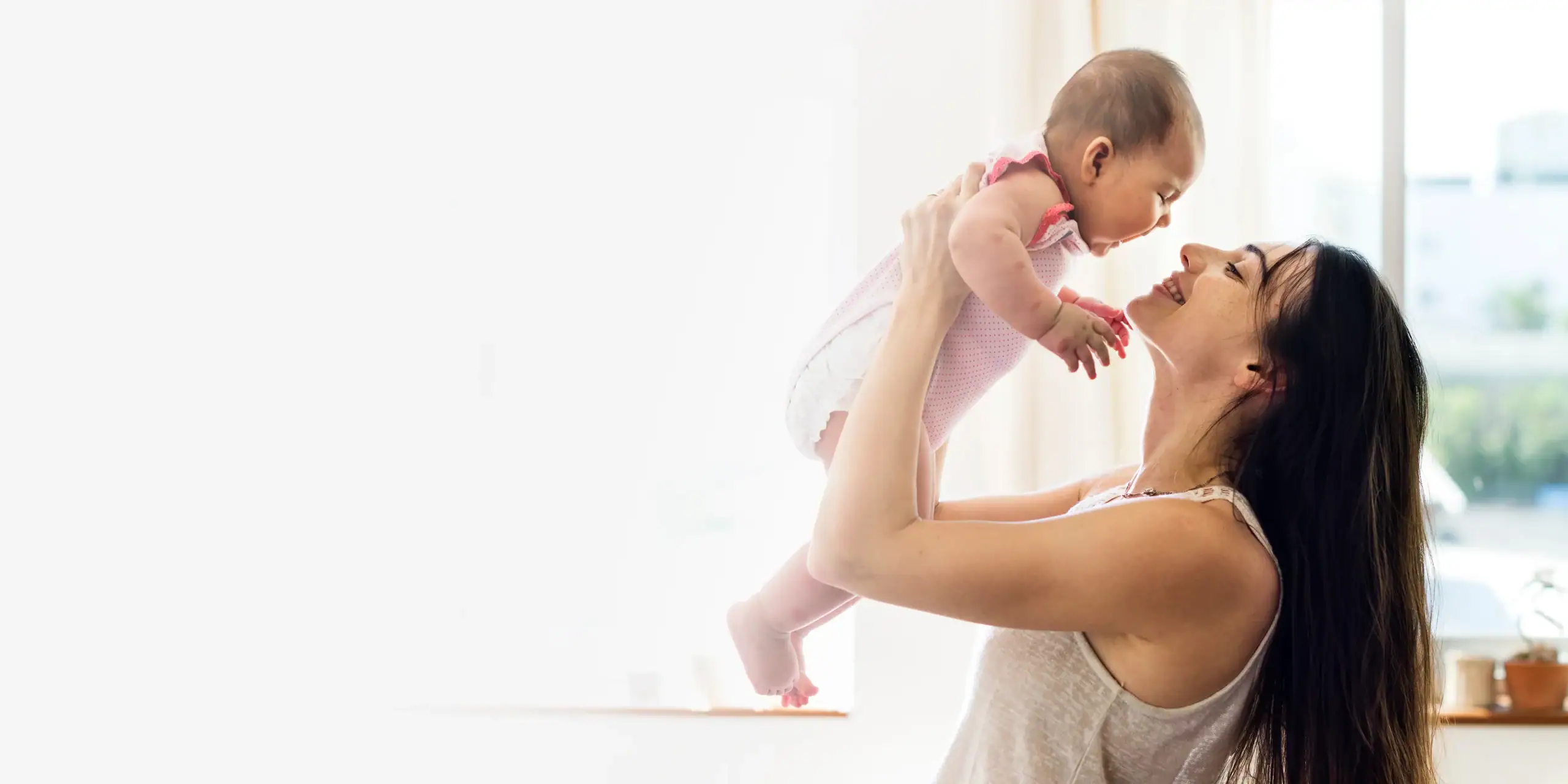 Postnatal Care in Dubai - A woman is holding a smiling baby up in the air near a bright window, looking lovingly at the child in a sunlit room.