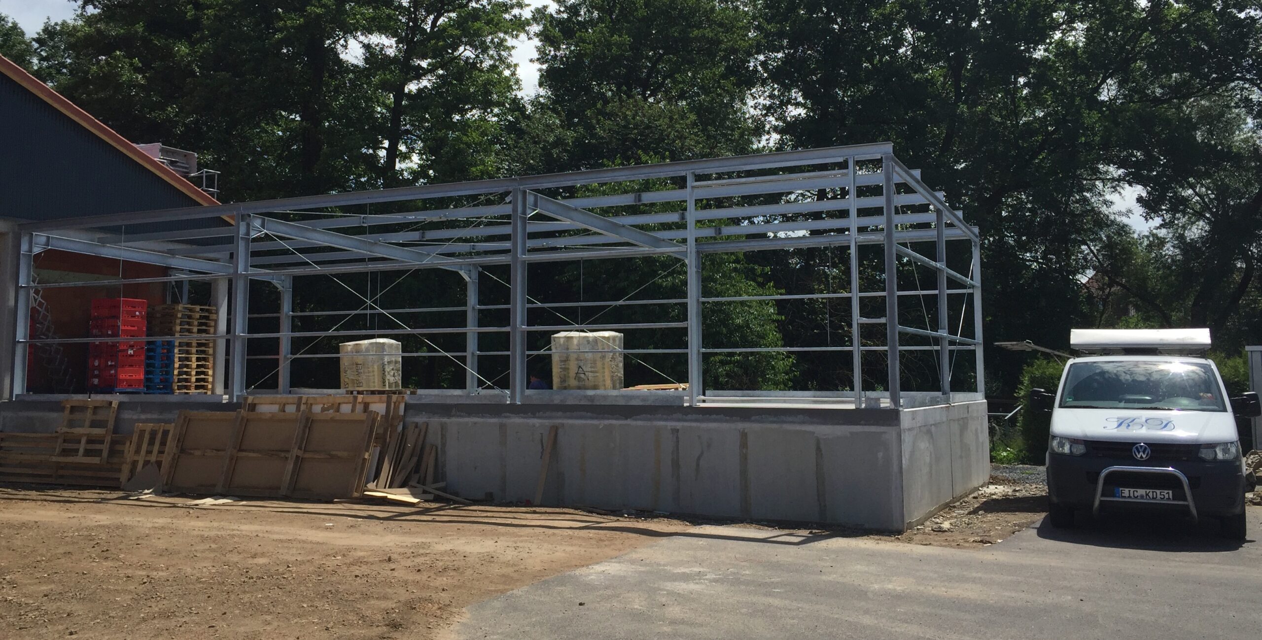Neubau-Stahlkonstruktion von Stahl- und Metallbau Drost. Das Bild zeigt einen Rohbau aus verzinktem Stahl mit Betonfundament, der als Anbau oder eigenständiges Gebäude dient, umgeben von Bäumen und einem Lieferwagen.