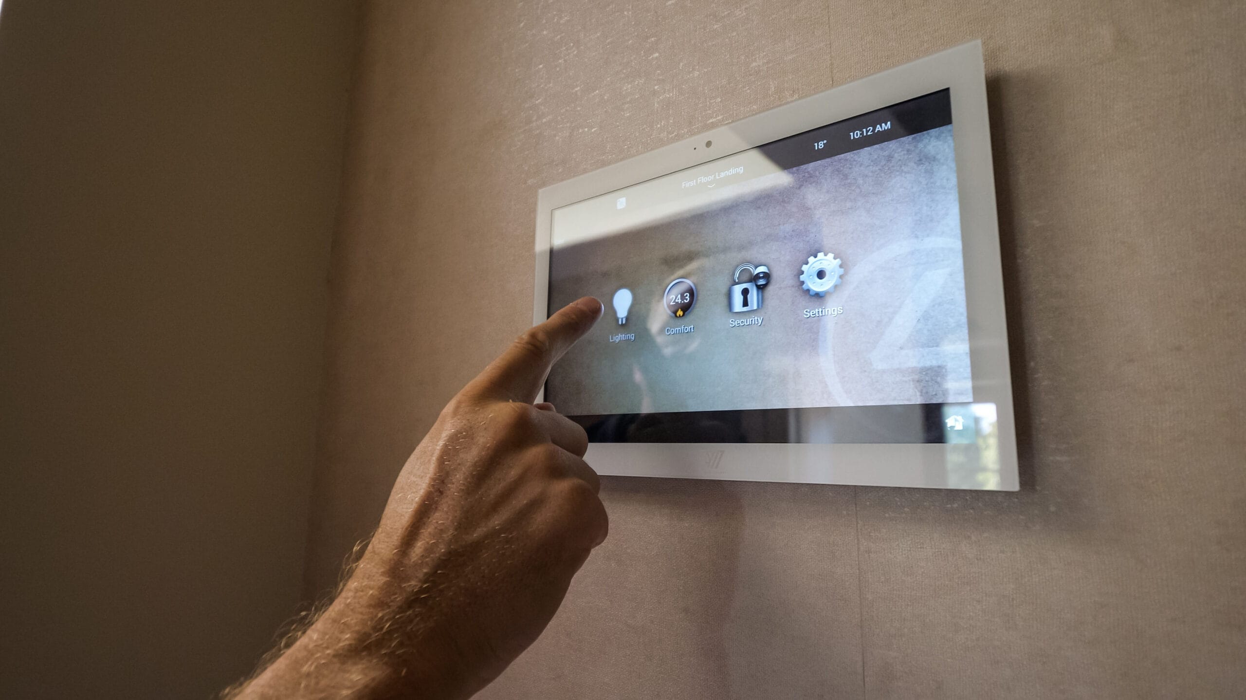 A hand is about to touch a wall-mounted touchscreen control panel displaying options for lighting, comfort, security, and settings—showcasing home automation installation in Surrey with a smart home interface.