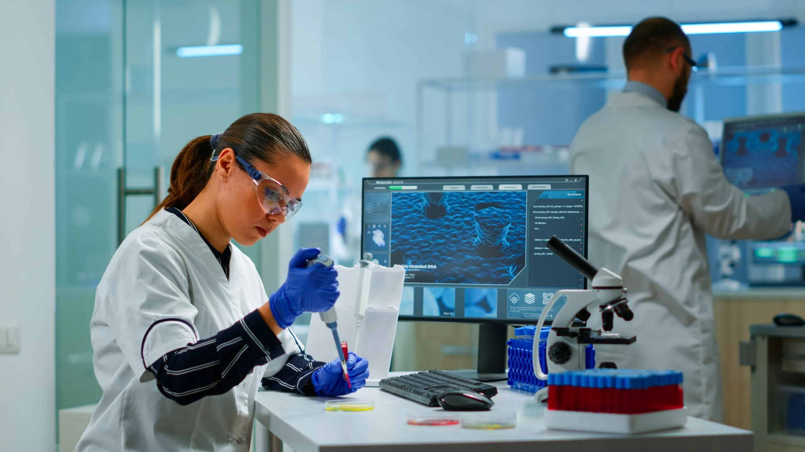 Scientist in a laboratory using a micropipette, surrounded by lab equipment and a computer displaying scientific data.
