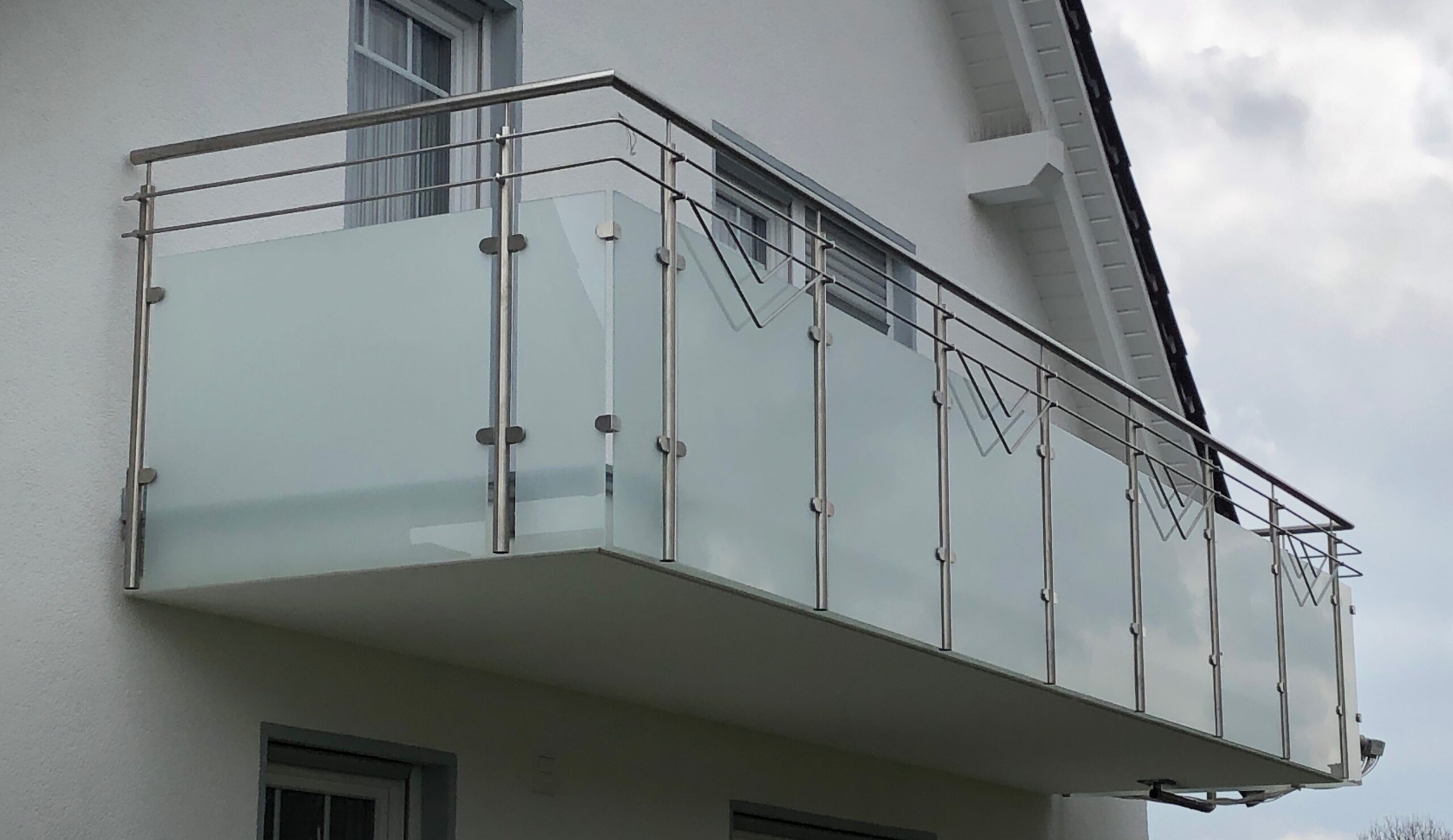 Modernes Balkongeländer von Stahl- und Metallbau Drost aus Edelstahl mit mattierten Glaspaneelen und Querstäben an einem weißen Haus.