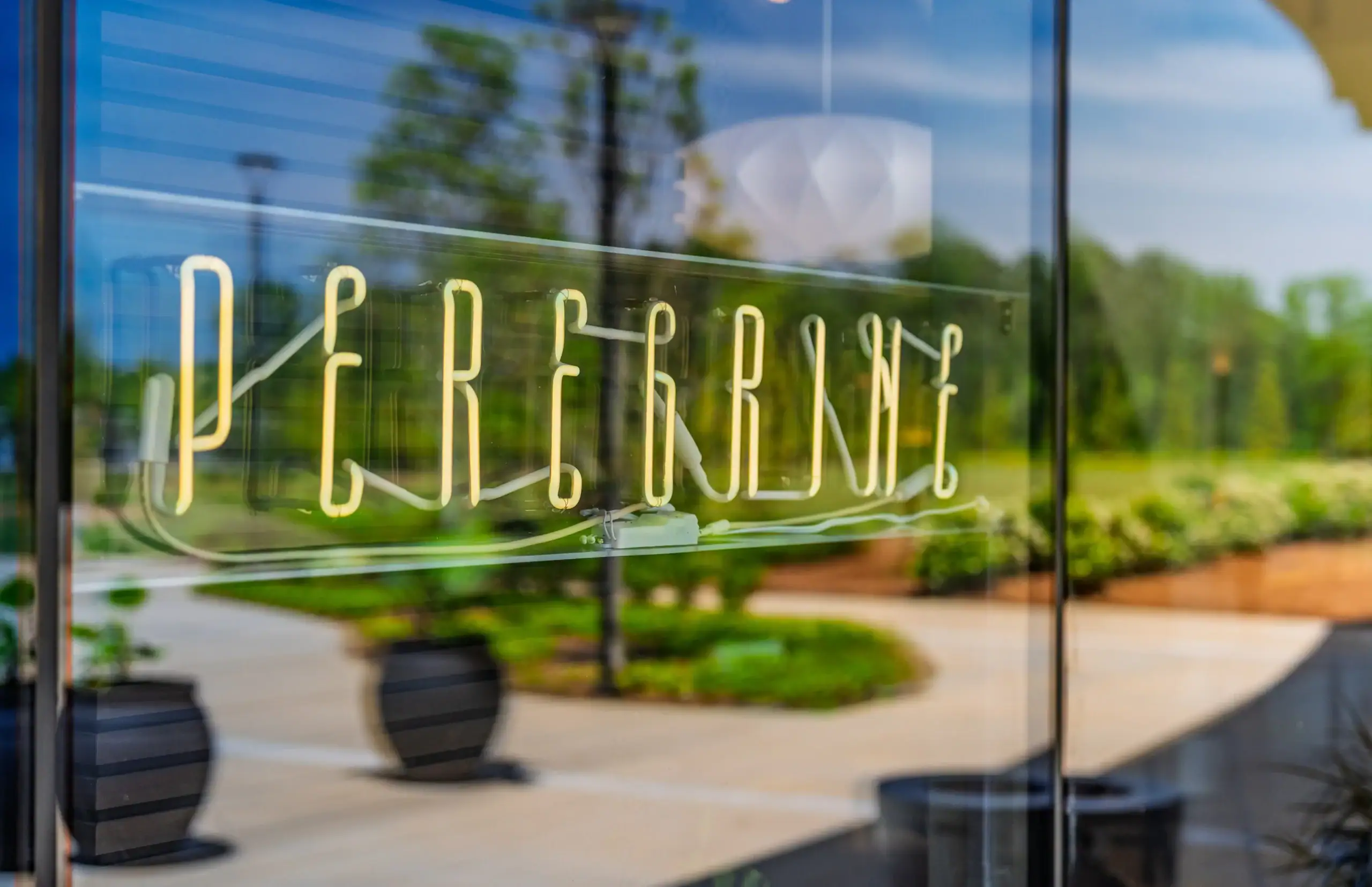 Illuminated neon sign for Peregrine, a Raleigh dining destination, displayed on a glass clubhouse entrance with landscaped grounds reflected in the modern façade.