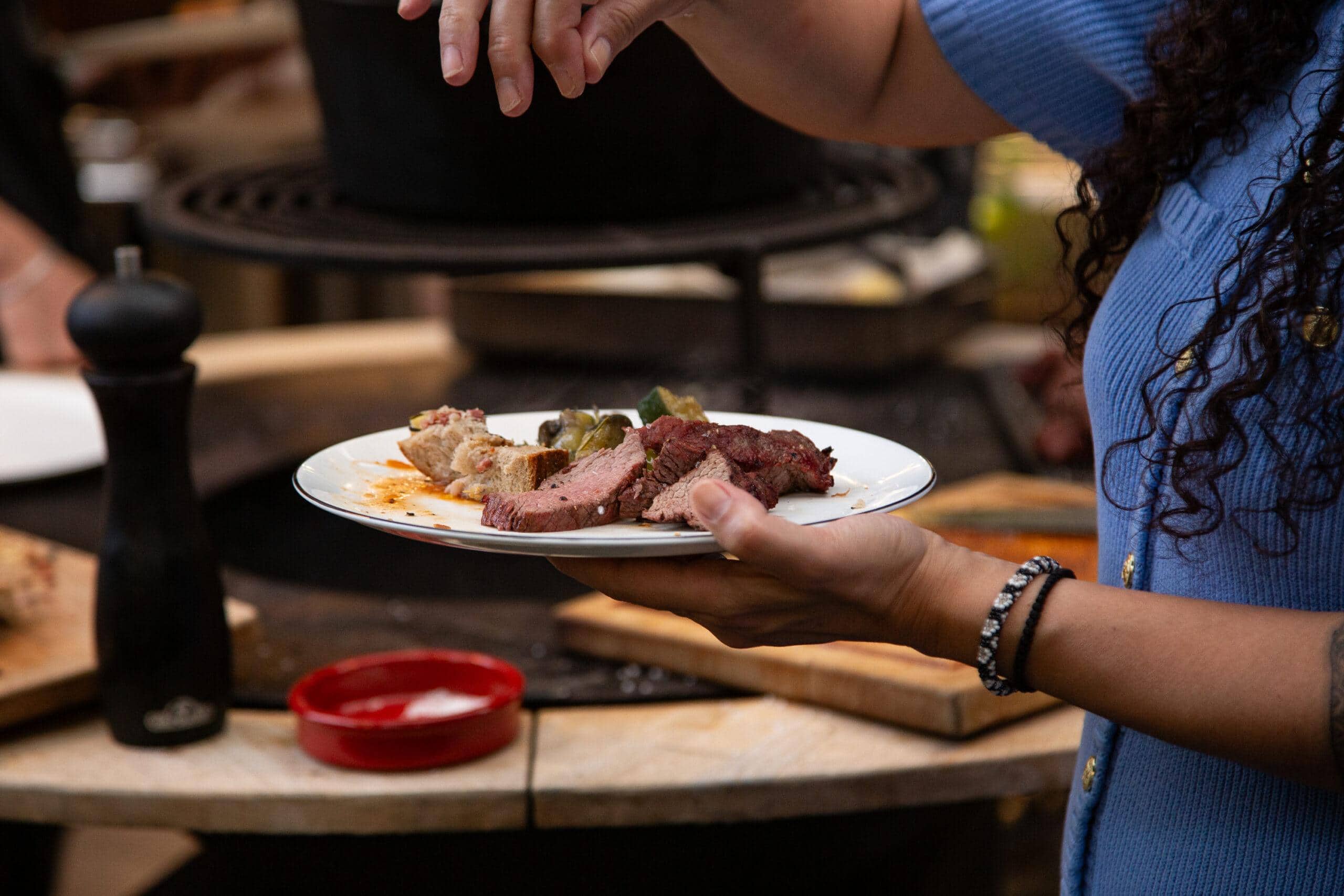 Fleisch angerichtet – Salz & Feuer im perfekten Moment Eine Frau hält im BBQ Raum Hürth einen Teller mit frisch zubereitetem Fleisch in der Hand und streut grobes Salz darüber – im Hintergrund unscharf der Puncher-Grill