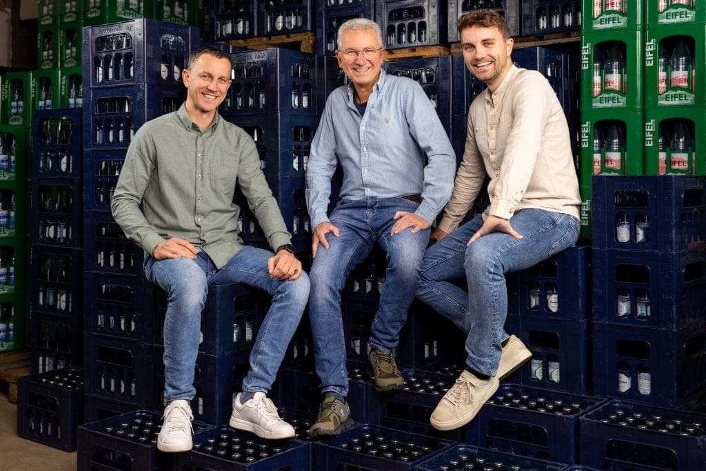 Drei Männer sitzen in einem Lagerraum mit Getränkekisten, lächelnd.