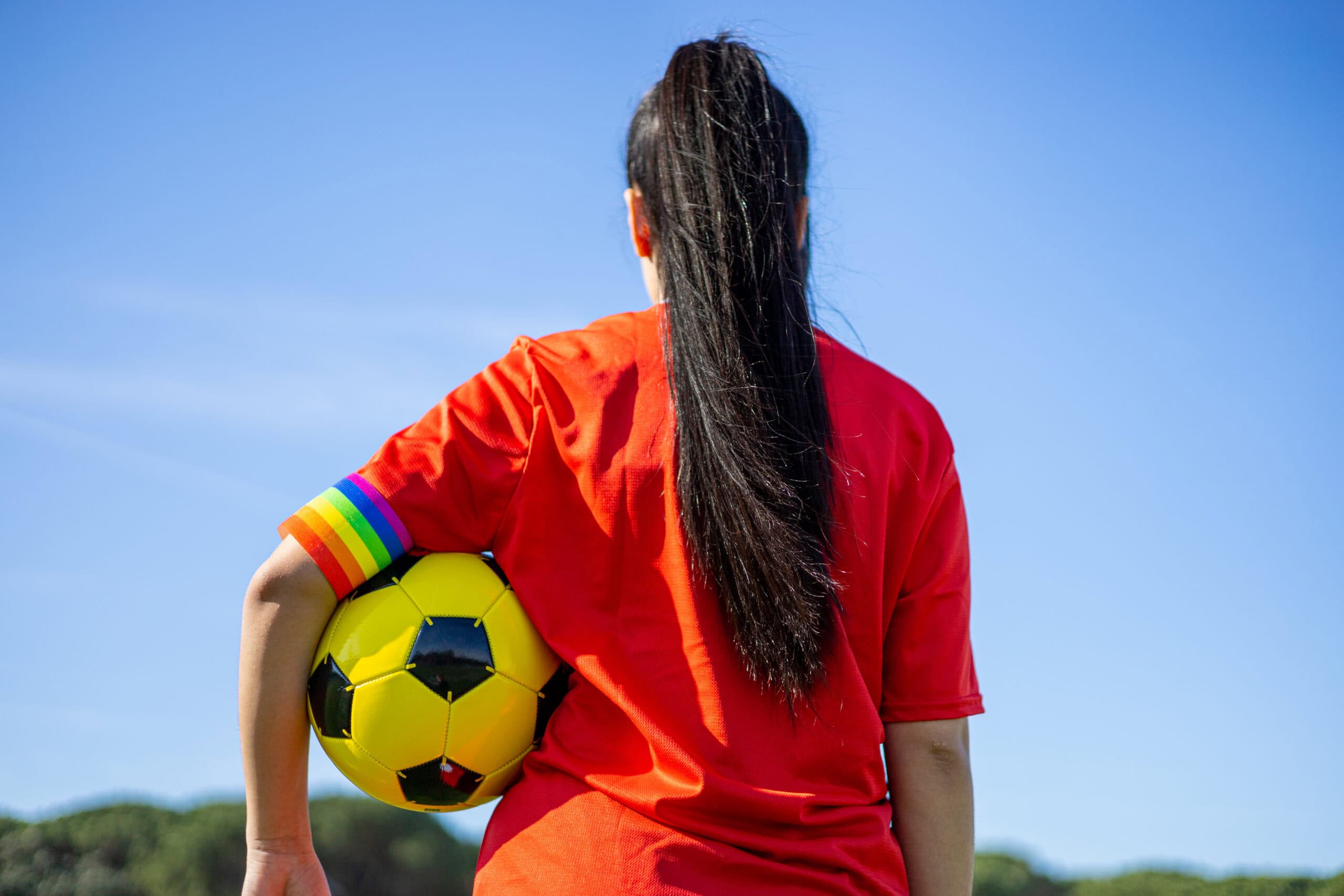 Realistic image of a back profile of a female athlete holding a yellow soccer ball with a rainbow band across her lower bicep.