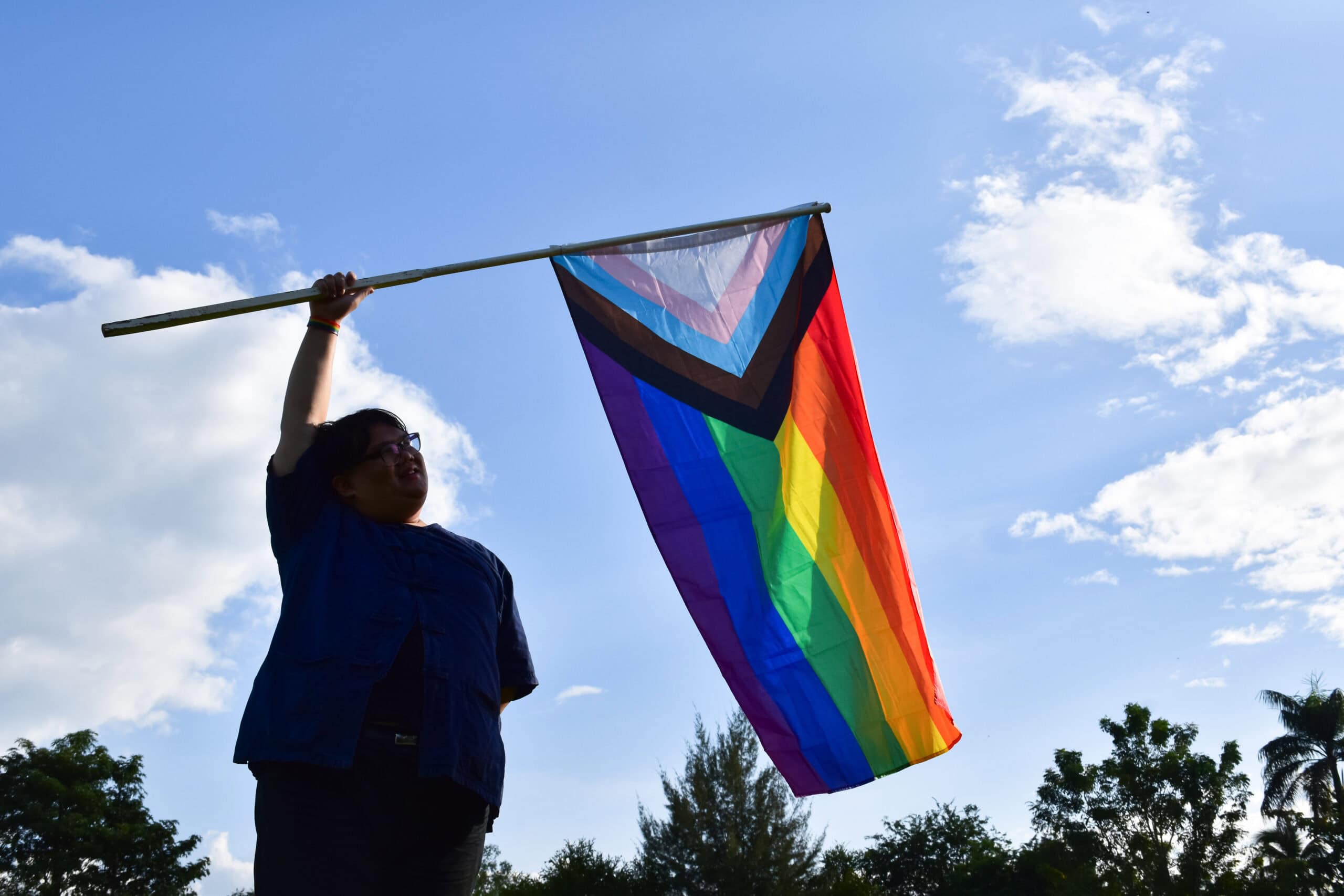 Realistic image of a woman holding an intersectional pride flag against the background of a blue sky with some whispy clouds.