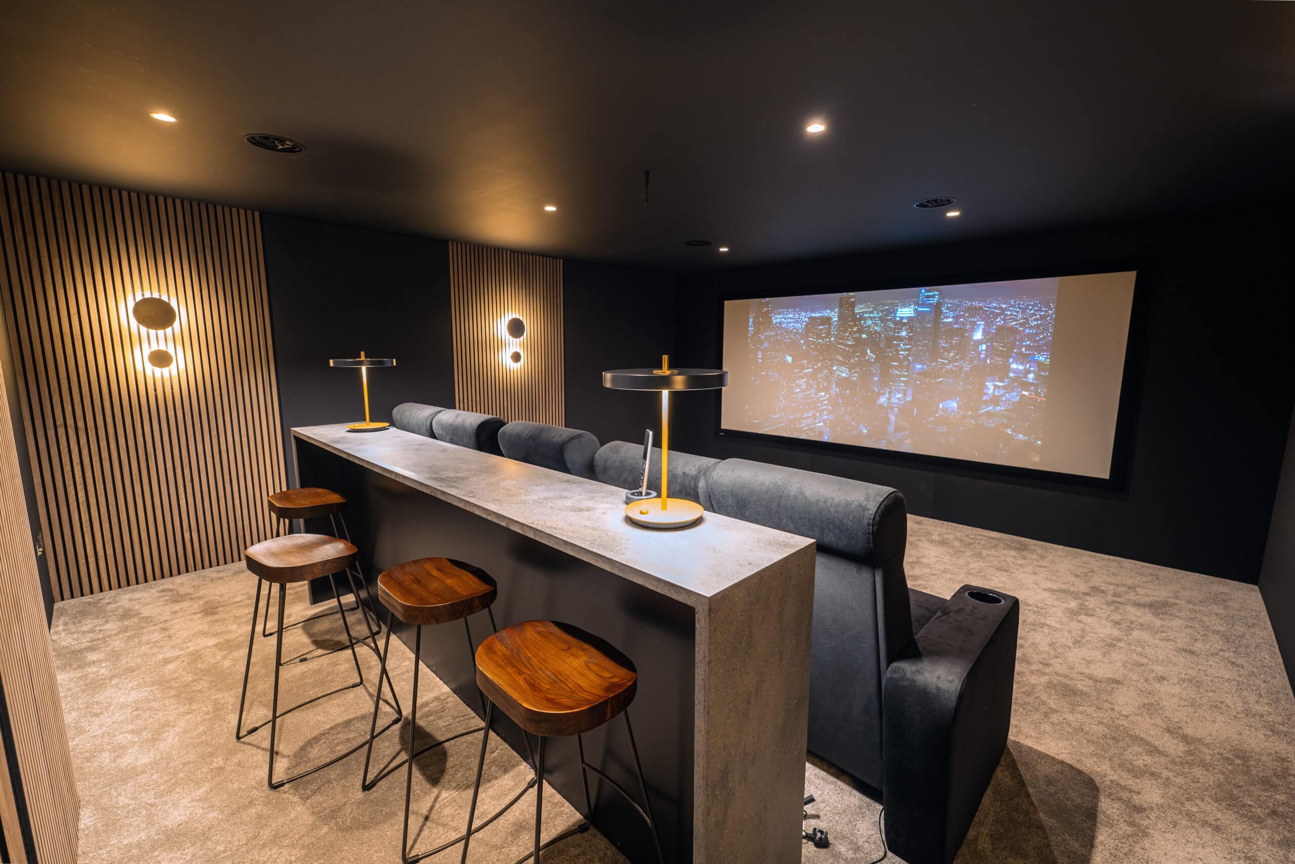 A modern home theatre room features a large screen showing a cityscape at night, plush recliner seats, and a high bar table with wooden stools—perfect for enjoying your home cinema with smart lighting installation in Virginia Waters, Surrey.