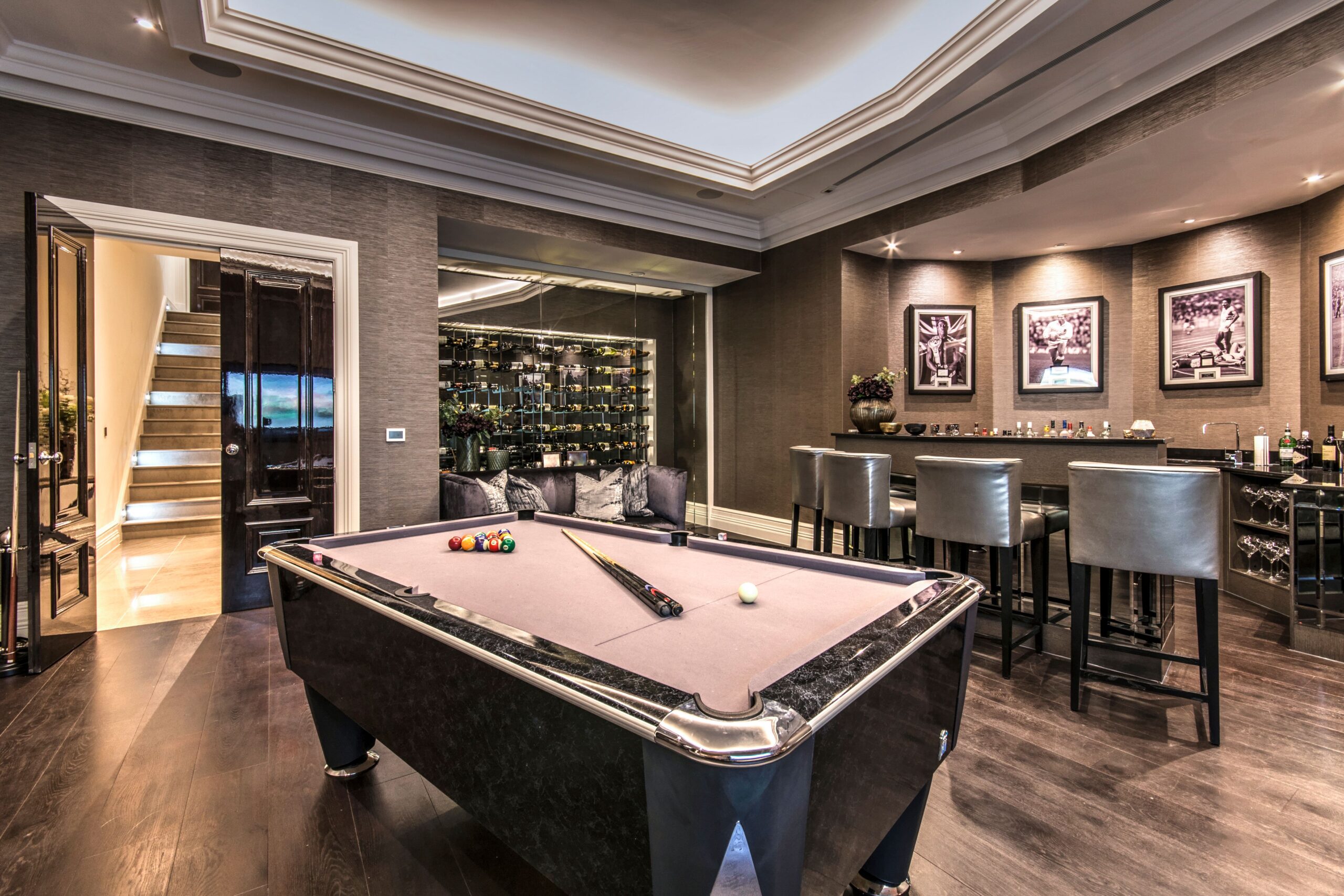 Luxurious games room with a pool table, modern bar area with high chairs, wine display, framed sports photos on the wall, and a staircase leading to a stunning basement home cinema with bar in Weybridge Surrey.
