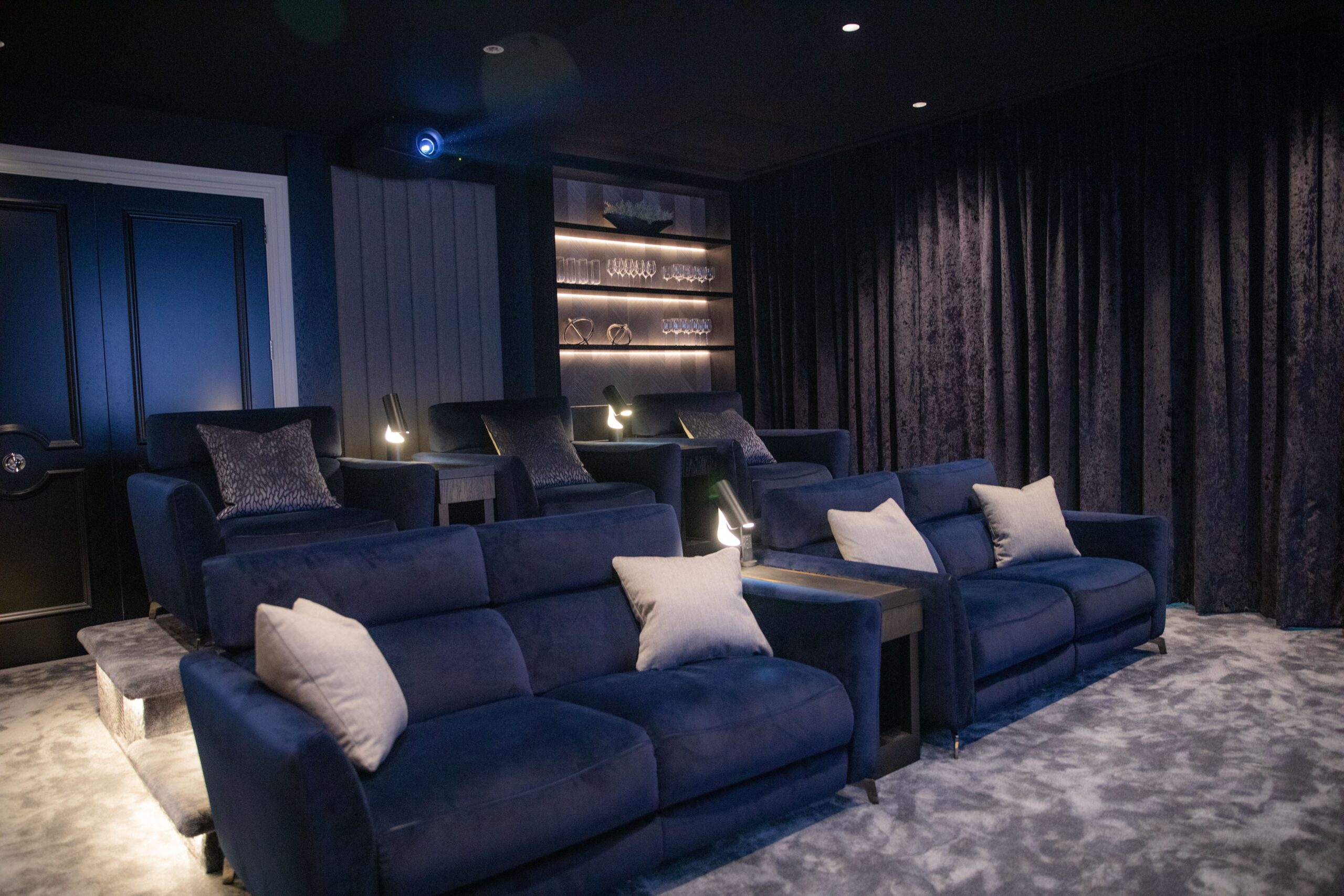 A luxurious home theatre room, showcasing a luxury home cinema installation in Walton-on-Thames, features tiered navy blue sofas, grey cushions, side tables, dim lighting, plush carpet, and shelves displaying glassware against a dark wall.