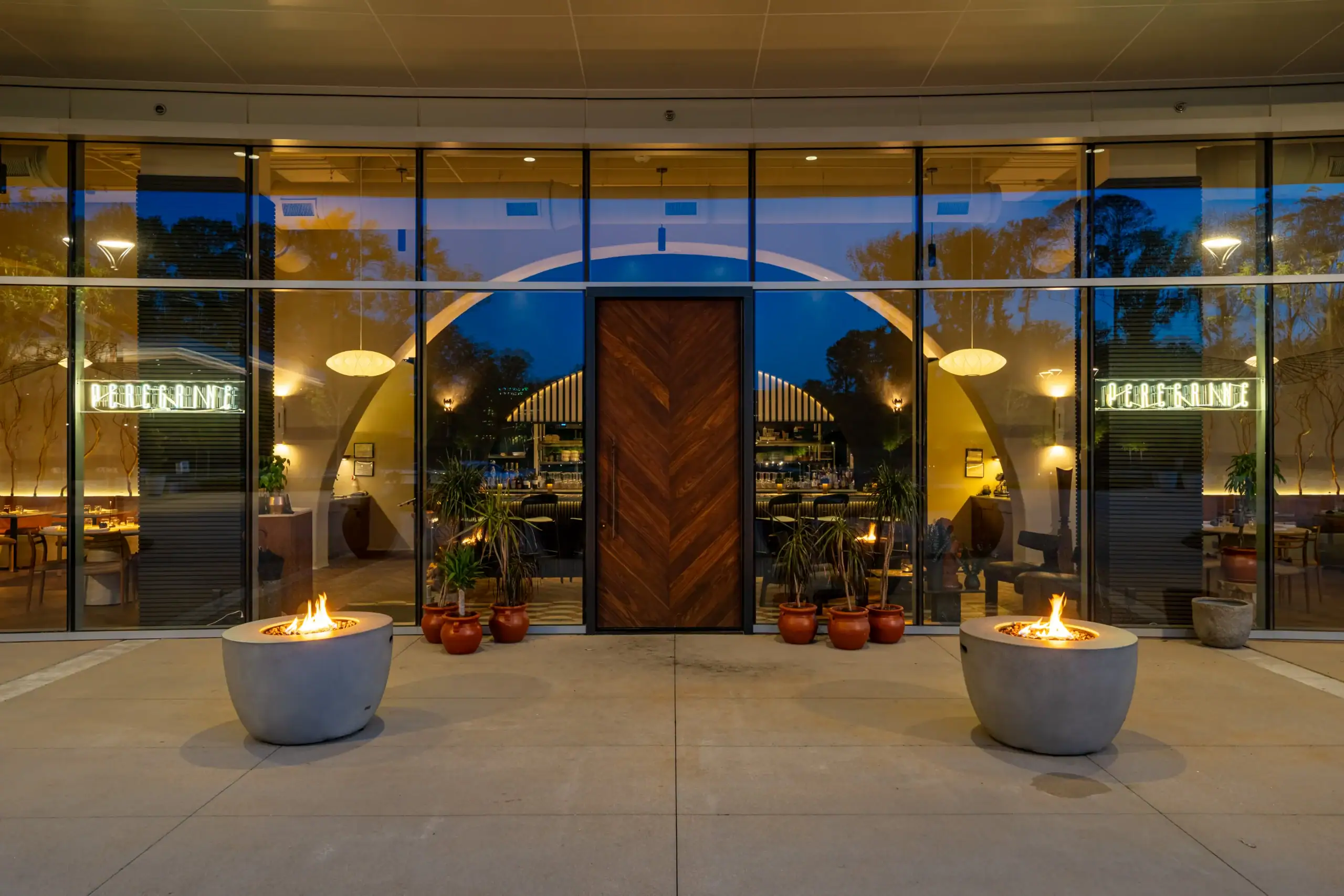 Elegant nighttime entrance to Peregrine, an upscale Raleigh restaurant featuring floor-to-ceiling glass, illuminated signage, a custom wood door, and outdoor fire pits.