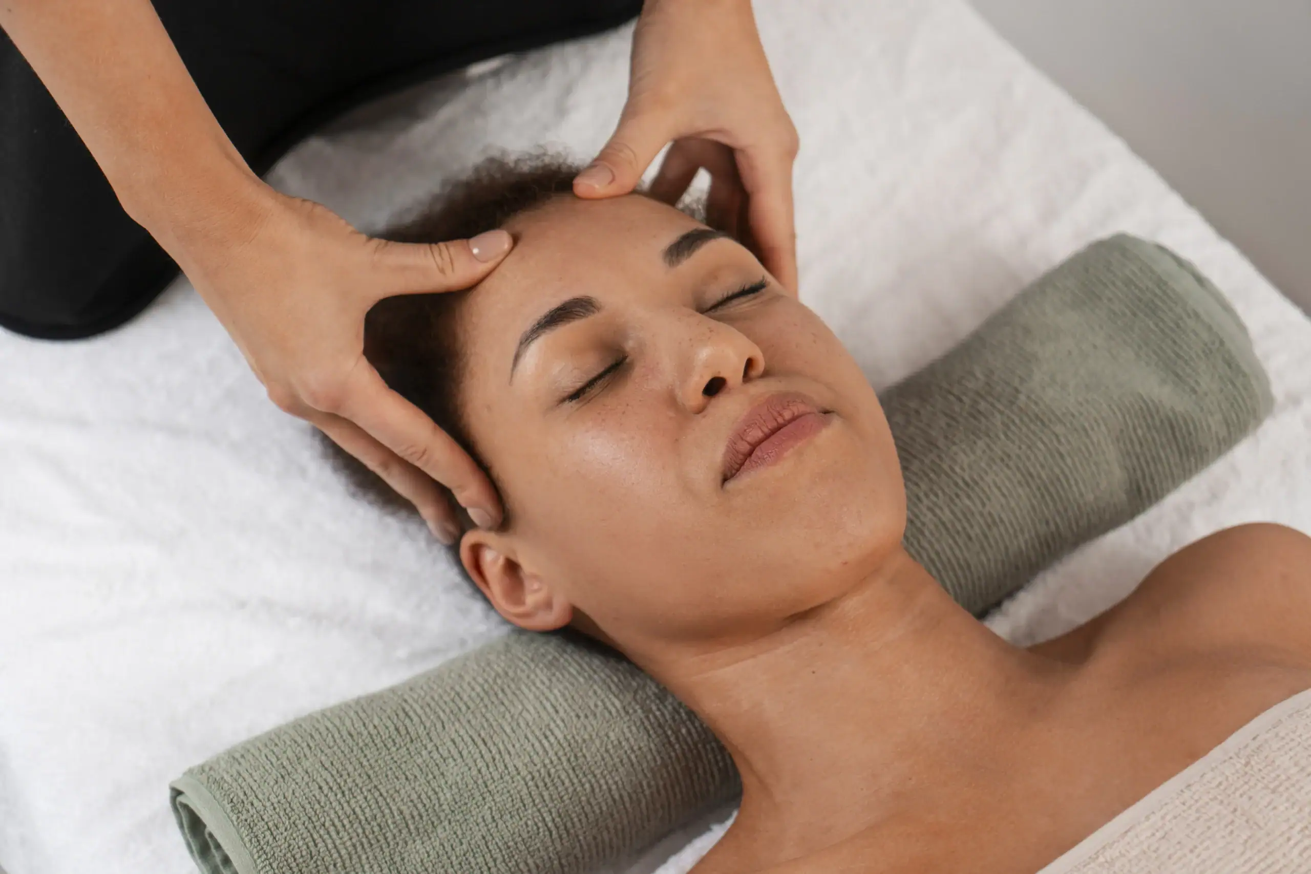Shiroabhyanga - An overhead view of a woman lying on her back with her eyes closed, receiving a head and temple massage. A practitioner’s hands are positioned on her forehead, applying gentle pressure. The woman’s head is supported by a rolled green towel on a white surface, conveying a sense of deep relaxation and professional care.