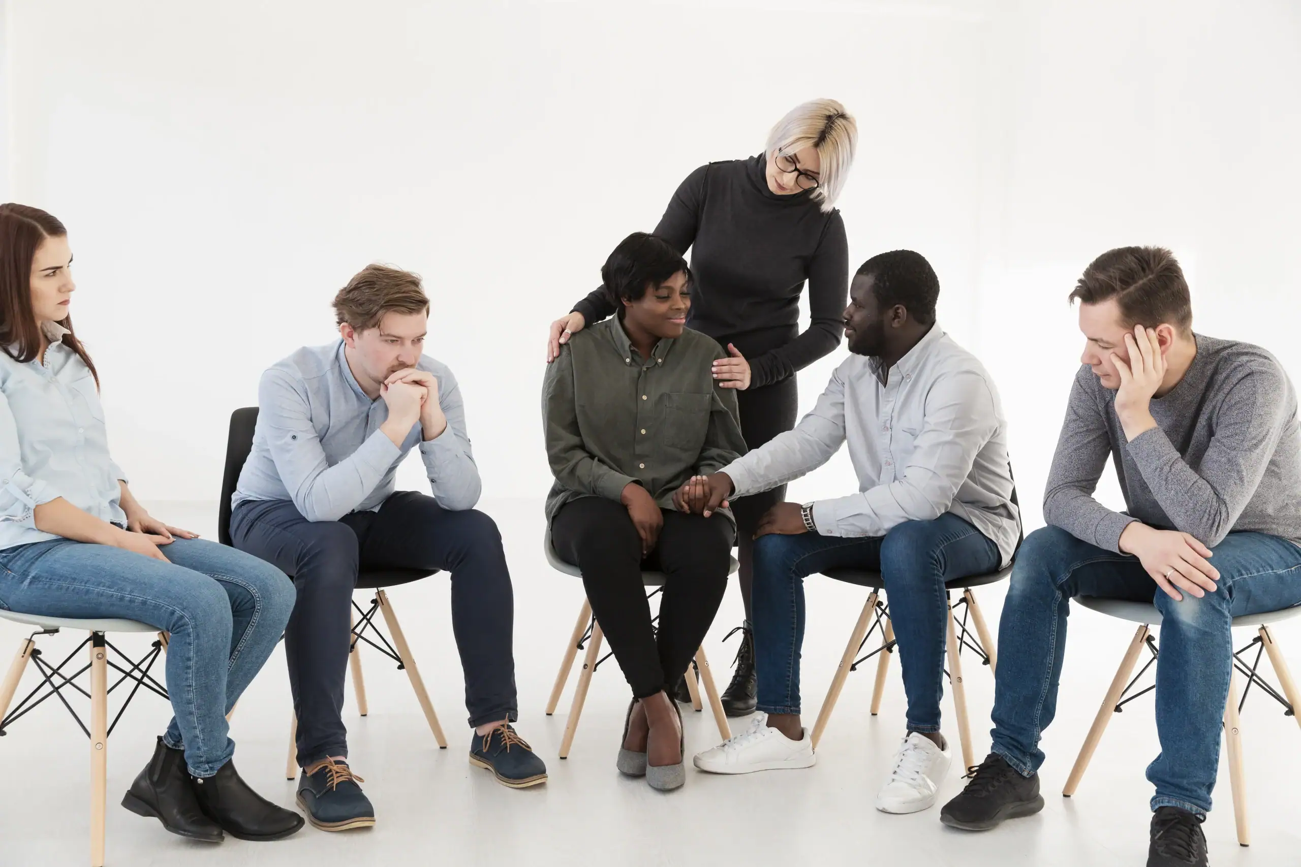 Addiction Recovery- In a bright, white setting, a diverse group of six people are gathered in a semi-circle, appearing to be in a supportive group setting.