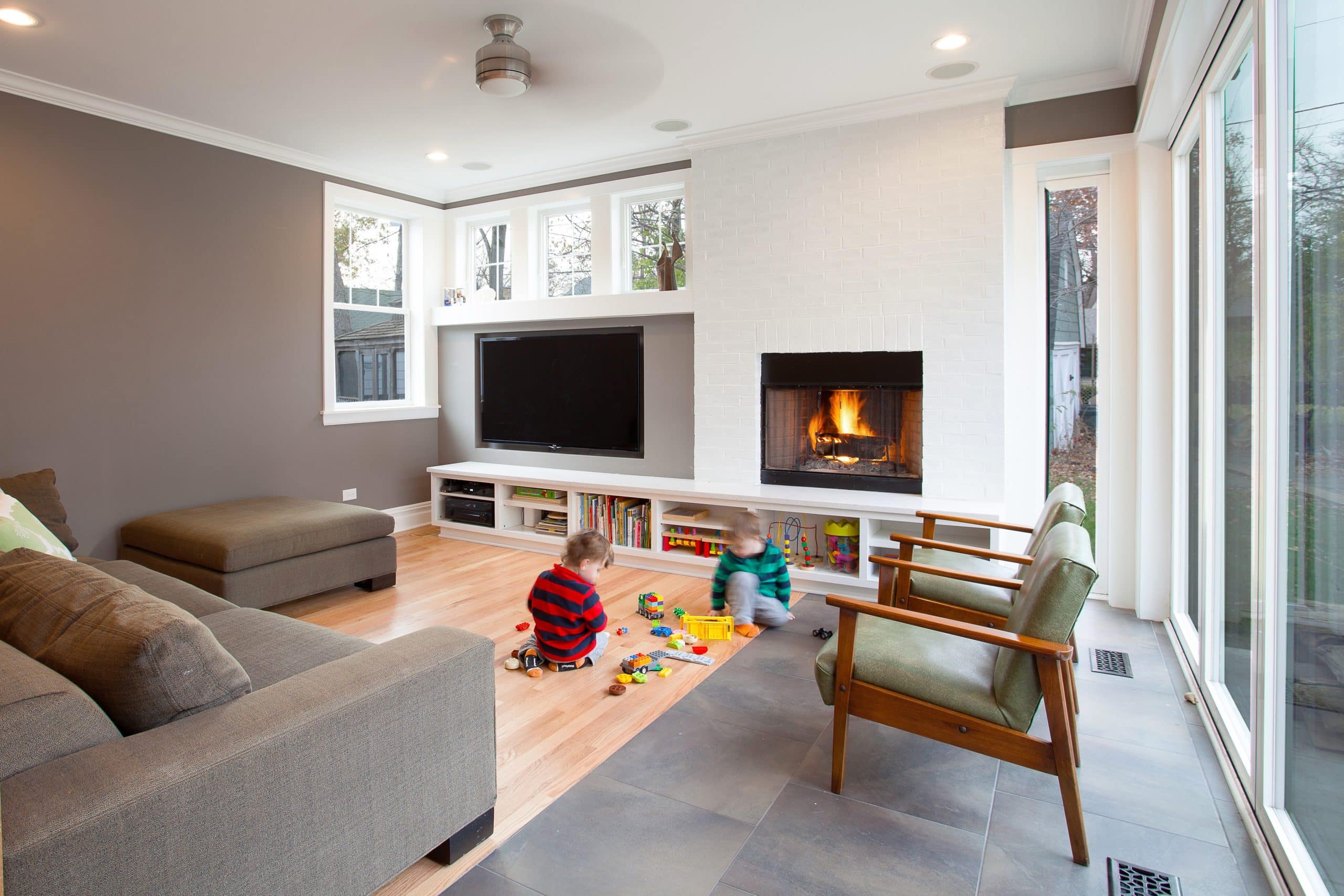 Two children playing in a living room with couch & chairs, built-in fireplace & mounted TV