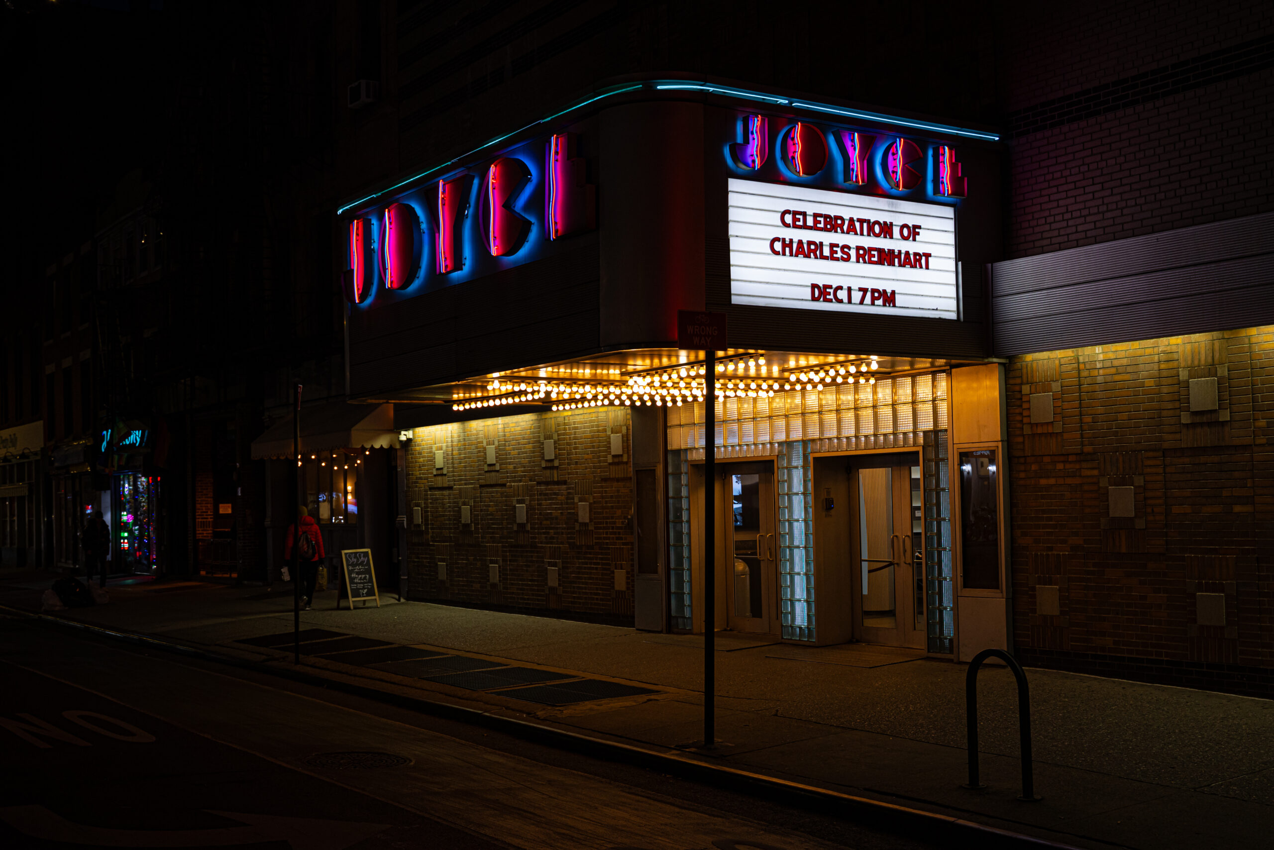 Night time photograph of the Joyce Theatre marquis with the name "Joyce" in pint and blue neon letters with the announcement "Celebration of Charles Reinhart, Dec 1, 7pm"