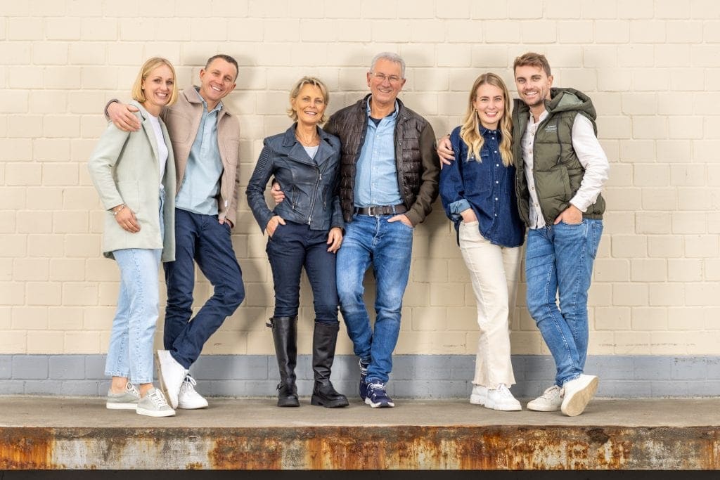 Gruppe von sechs Menschen, die vor einer Wand stehen, lächelnd und freundlich.