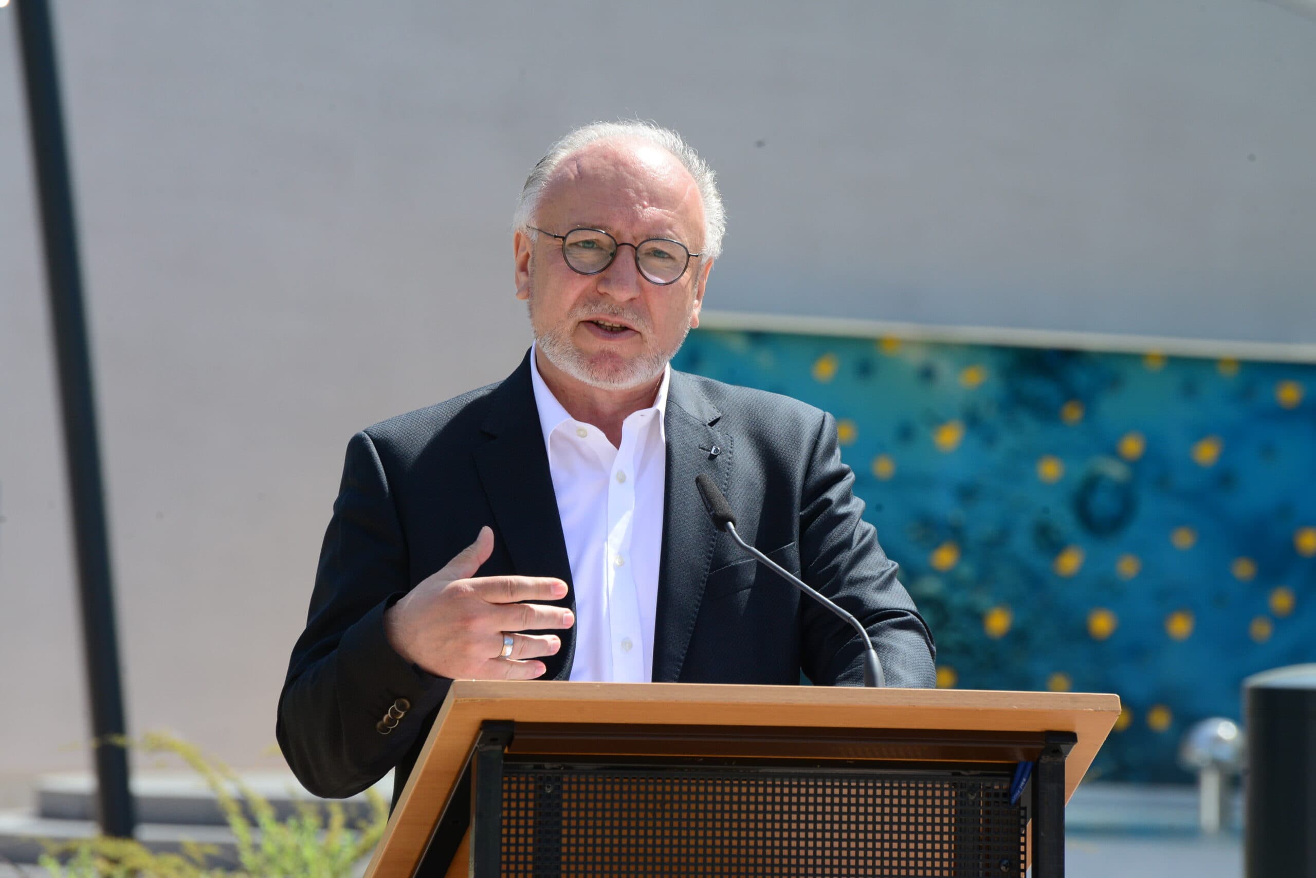 Ralph Hauboldt steht im Vordergrund, an einem Podium und hält eine Rede.