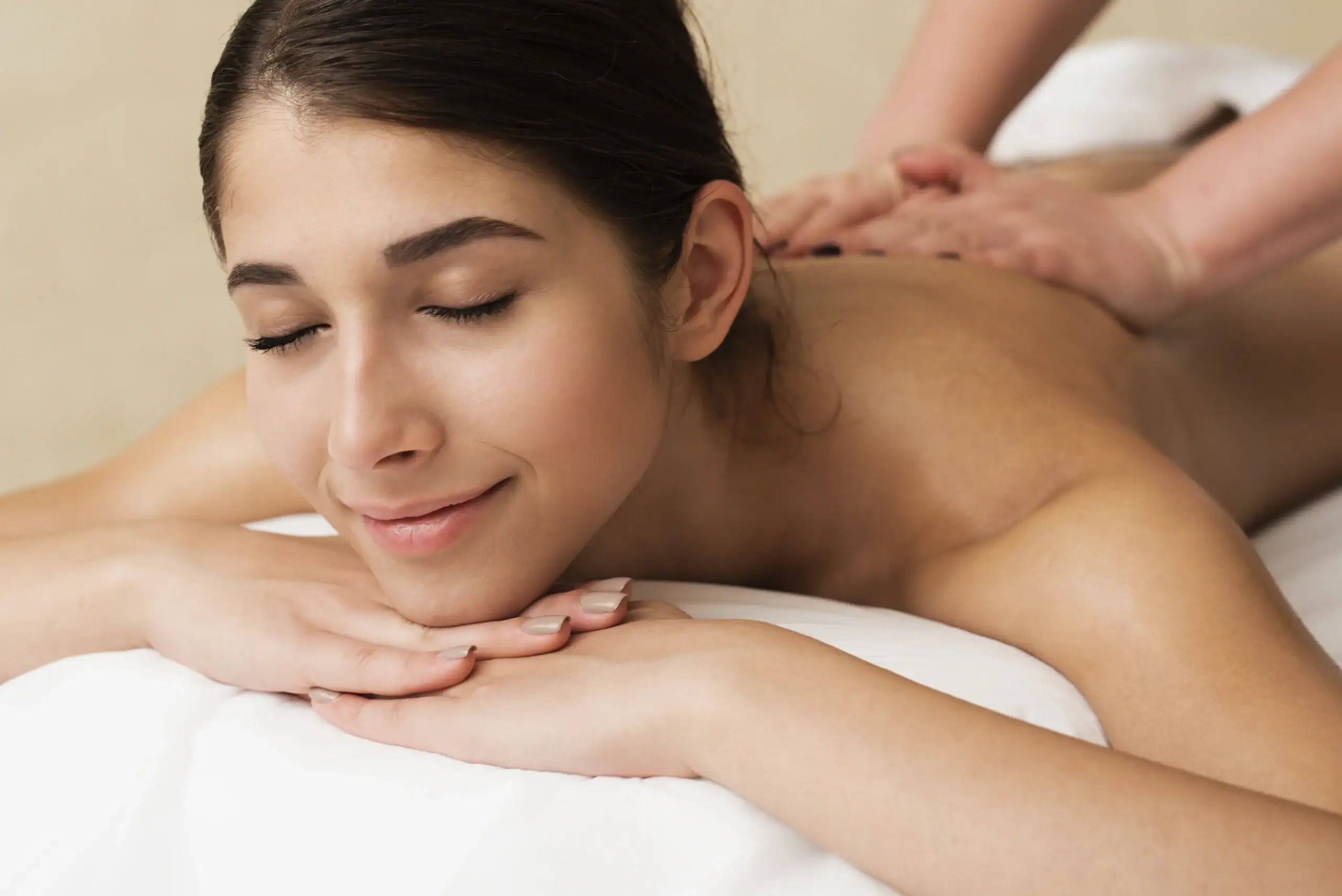 Abhyanga- A close-up of a young woman with her eyes closed and a peaceful smile, lying on her stomach while receiving a back massage.