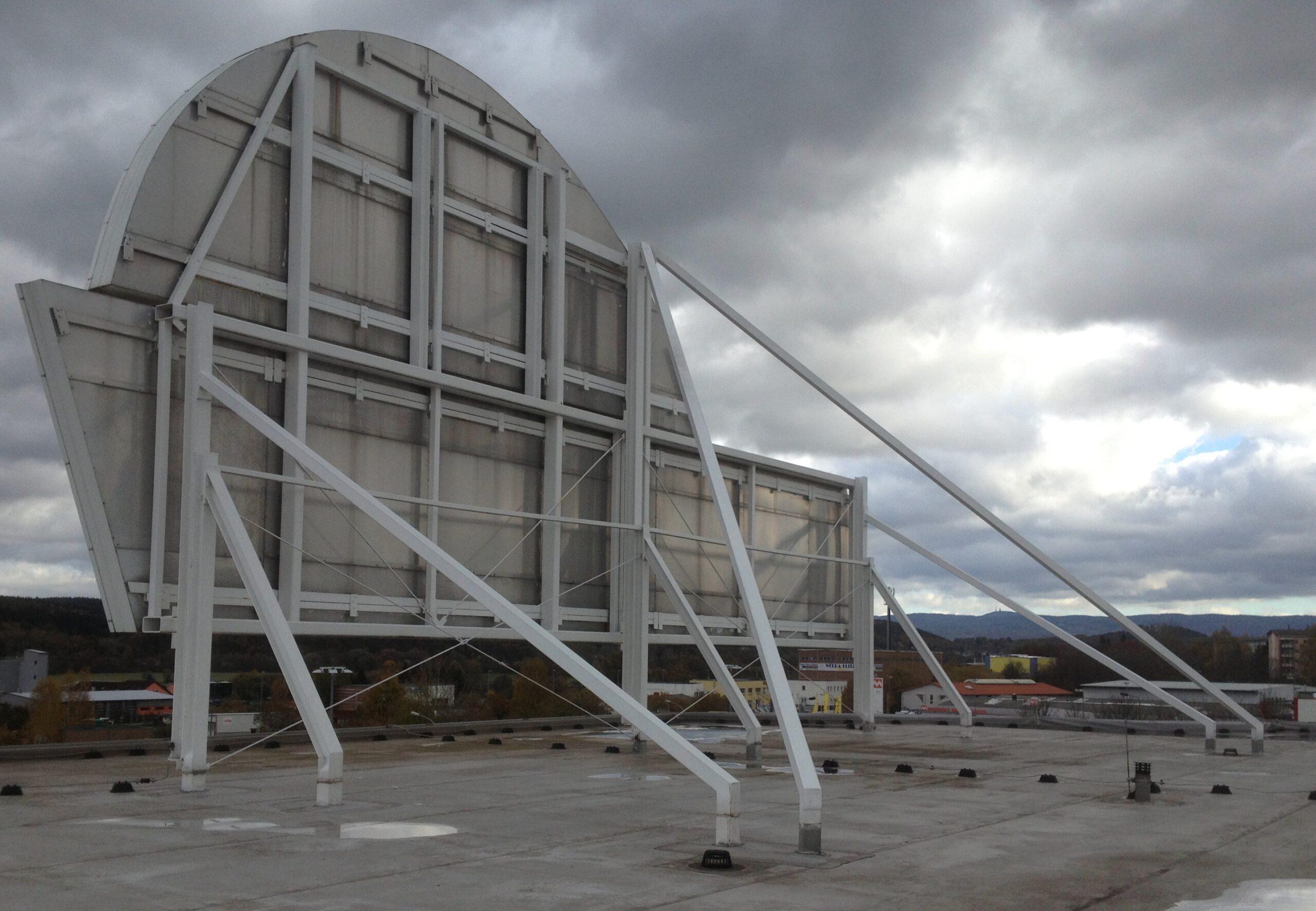 Rückansicht einer großen, gebogenen Anzeigetafel aus weißem Stahl, fachmännisch konstruiert von Stahlbau von Stahl- und Metallbau Drost, auf einem Flachdach. Die robuste Stahlkonstruktion ist mit zahlreichen Stützen und Verstrebungen versehen, die ihre Stabilität unter einem bewölkten Himmel zeigen. Im Hintergrund sind bewaldete Hügel und ferne Gebäude zu erkennen.
