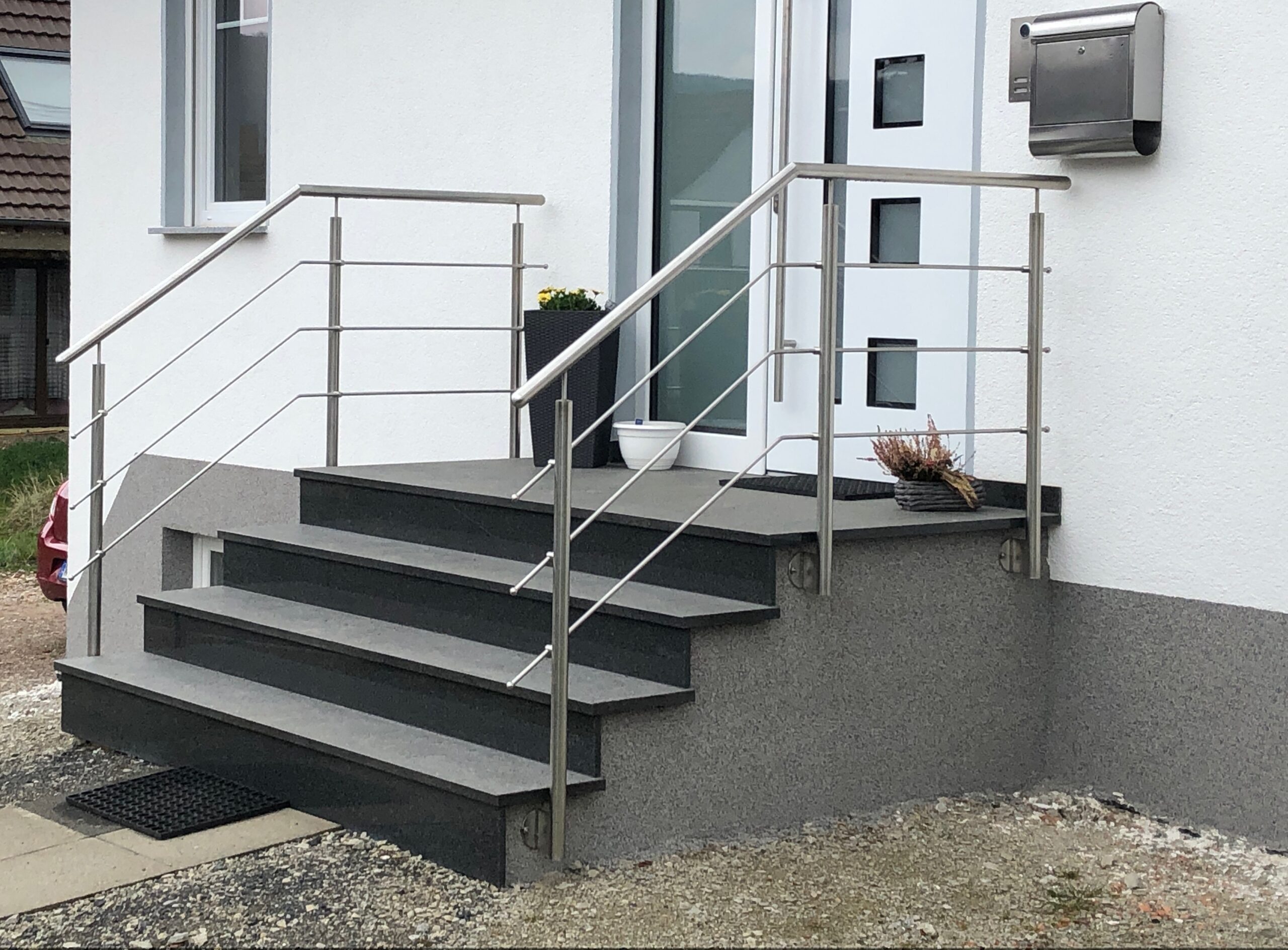 Ein modernes Edelstahlgeländer von Stahl- und Metallbau Drost an einer Außentreppe mit dunklen Stufen, die zu einem weißen Hauseingang führen.