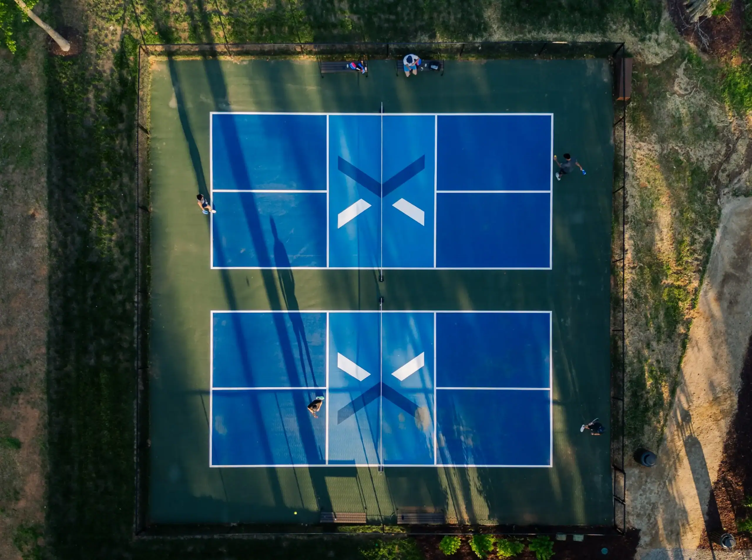 Aerial view of modern outdoor pickleball courts with players enjoying matches in a scenic community surrounded by trees and green space.