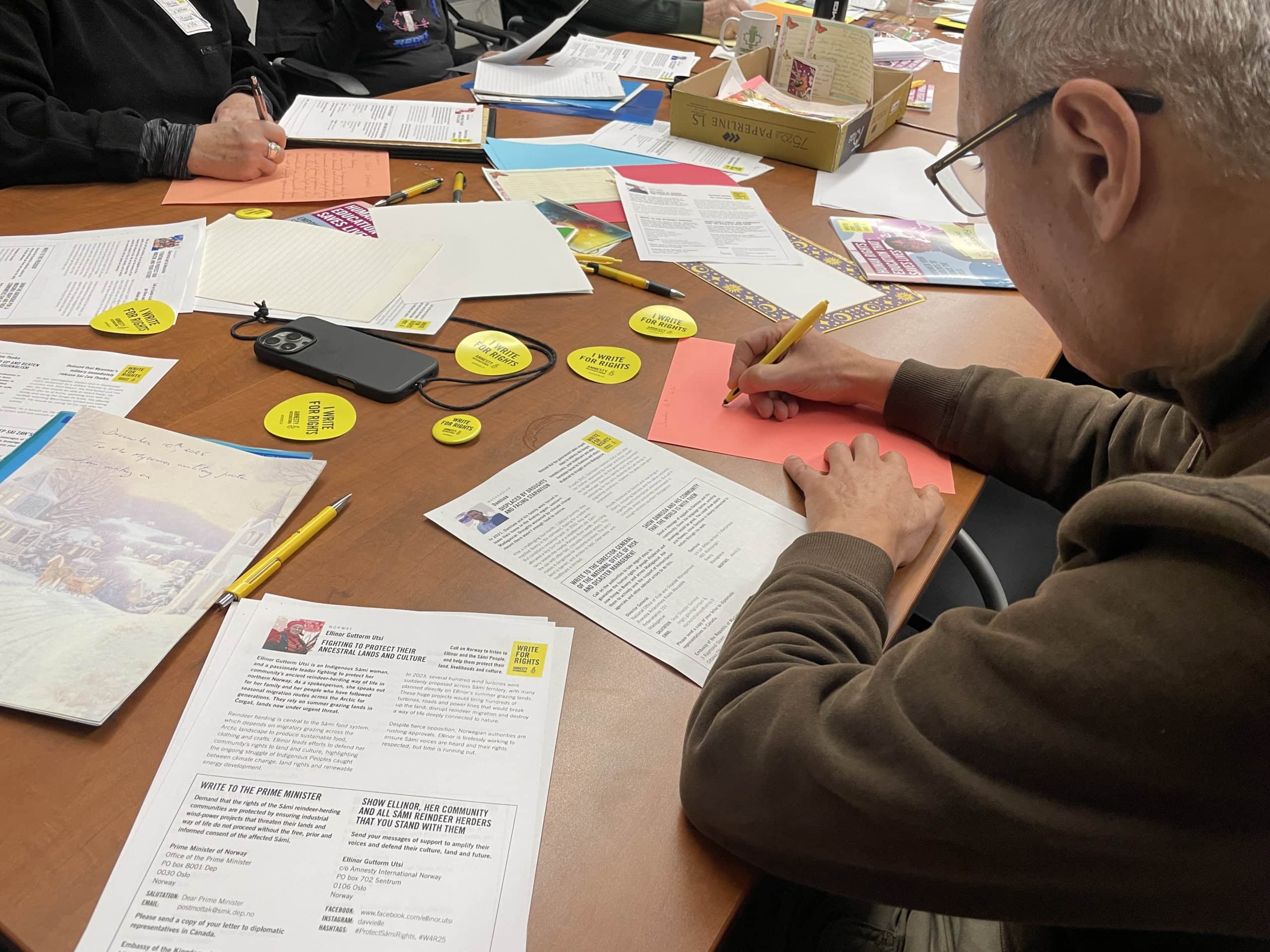 “Amnesty International supporters writing handwritten letters at a table covered with case sheets, yellow pens, and Write for Rights materials.”