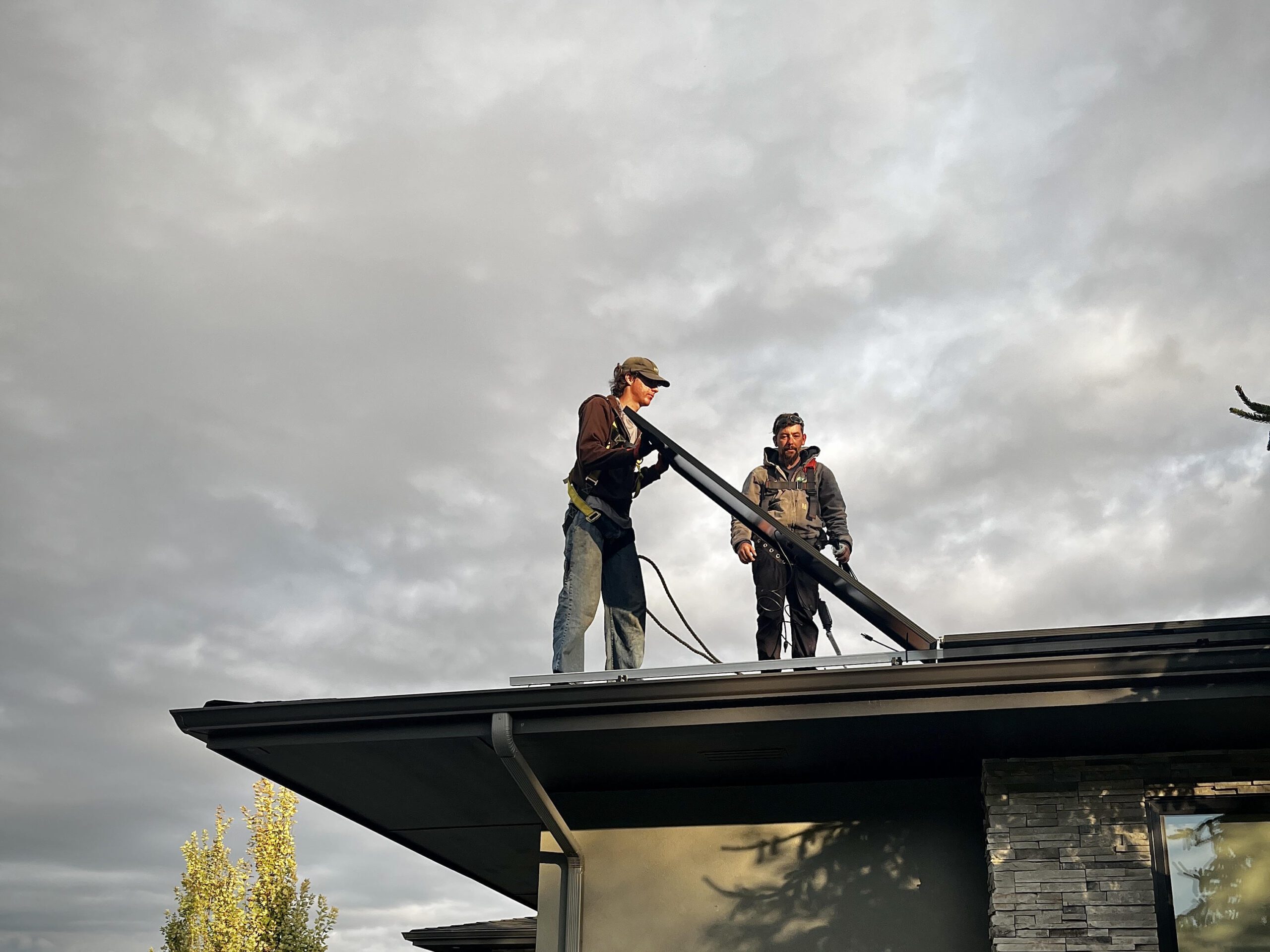Two individuals on a roof, and one is holding a solar panel. The sky is cloudy in the background.