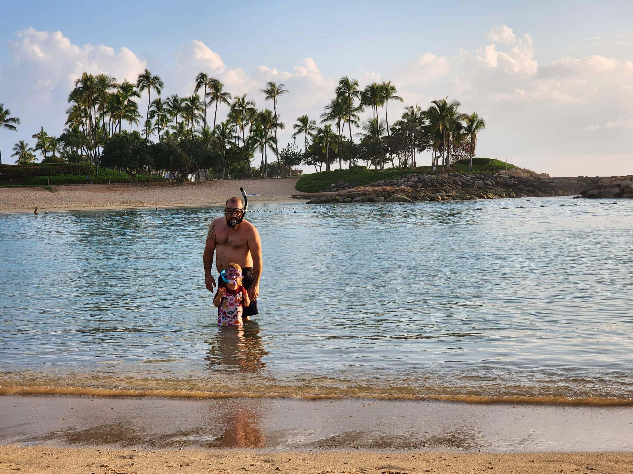 Justin and River enjoying the warm water at a serene beach in Kapolei, Oahu. Both are wearing snorkel gear, with lush palm trees and a sandy shoreline creating a tropical backdrop.