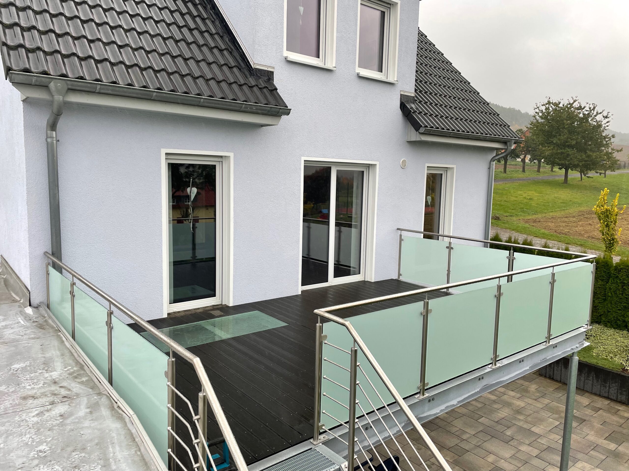 Moderner Balkon von Stahl- und Metallbau Drost mit dunkelbraunem Holzdeck, Edelstahlgeländer und mattierter Glasbrüstung, inklusive Außentreppe. Ein transparentes Glasbodenelement ist im Deck integriert.
