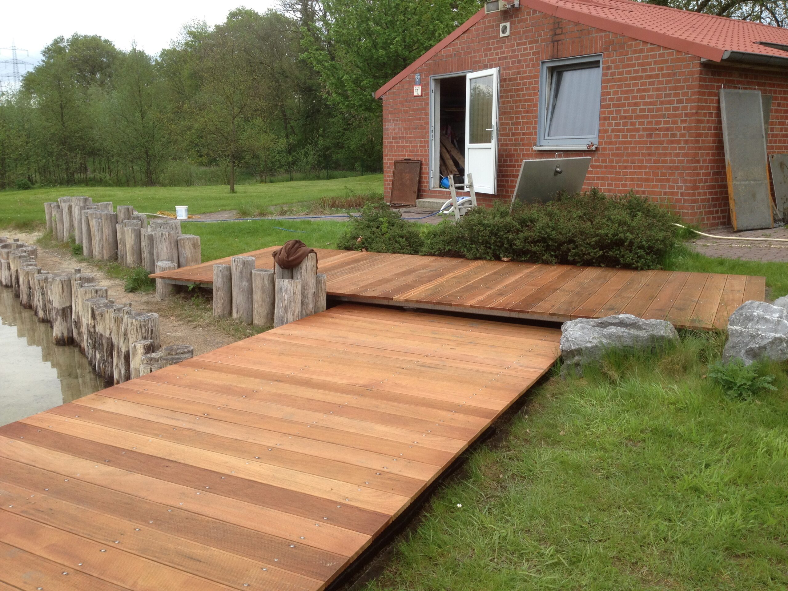 Hochwertiger, maßgefertigter Holzsteg von Sonderanfertigungen von Stahl- und Metallbau Drost, der von einer Rasenfläche an ein Gewässer grenzt und von rustikalen Holzpfählen gesäumt wird. Im Hintergrund ist ein rotes Backsteingebäude und Bäume zu sehen.