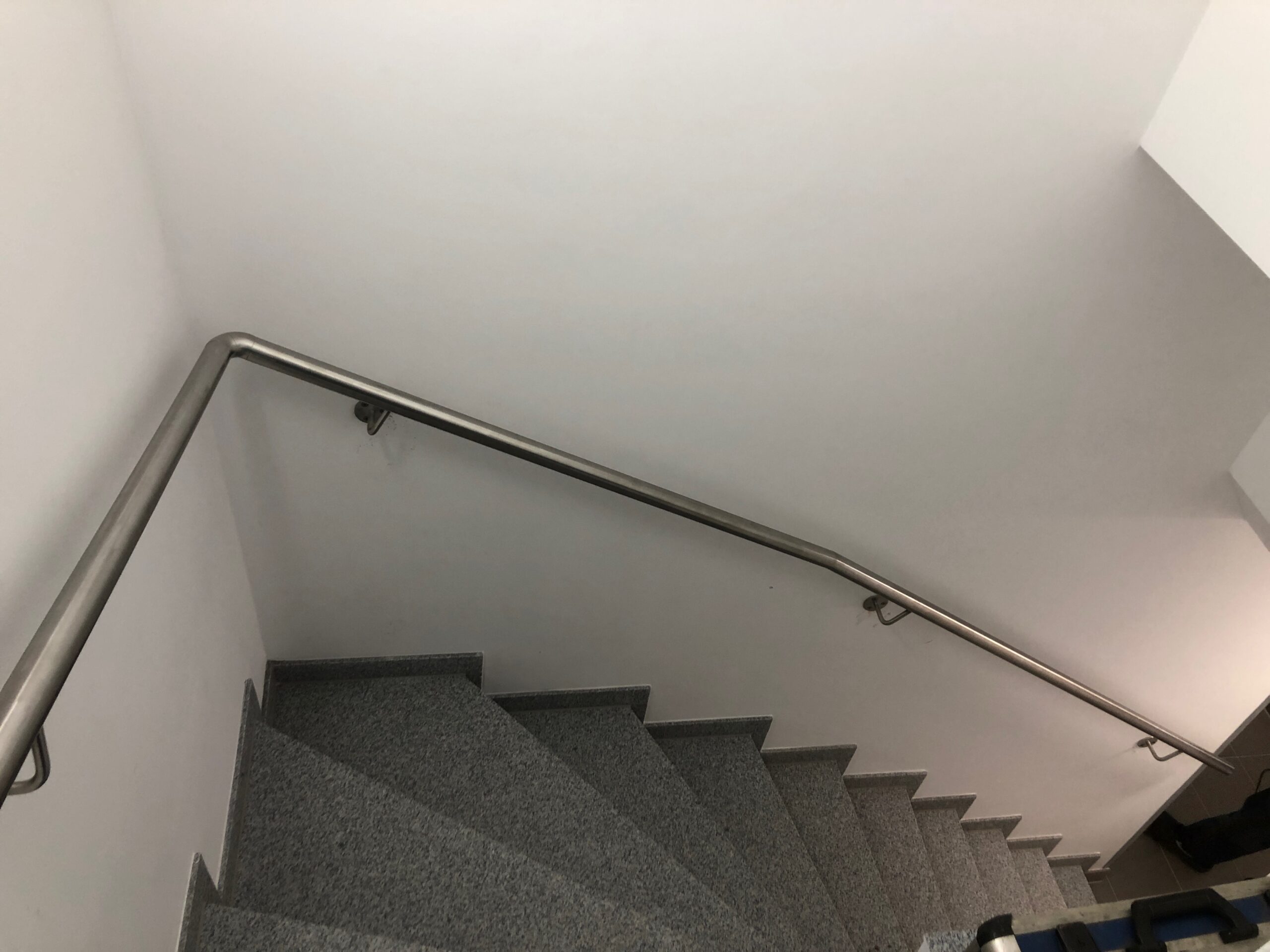 Ein moderner, gebogener Edelstahl-Handlauf von Stahl- und Metallbau Drost ist an einer hellen Wand entlang einer dunkelgrauen Granittreppe montiert.