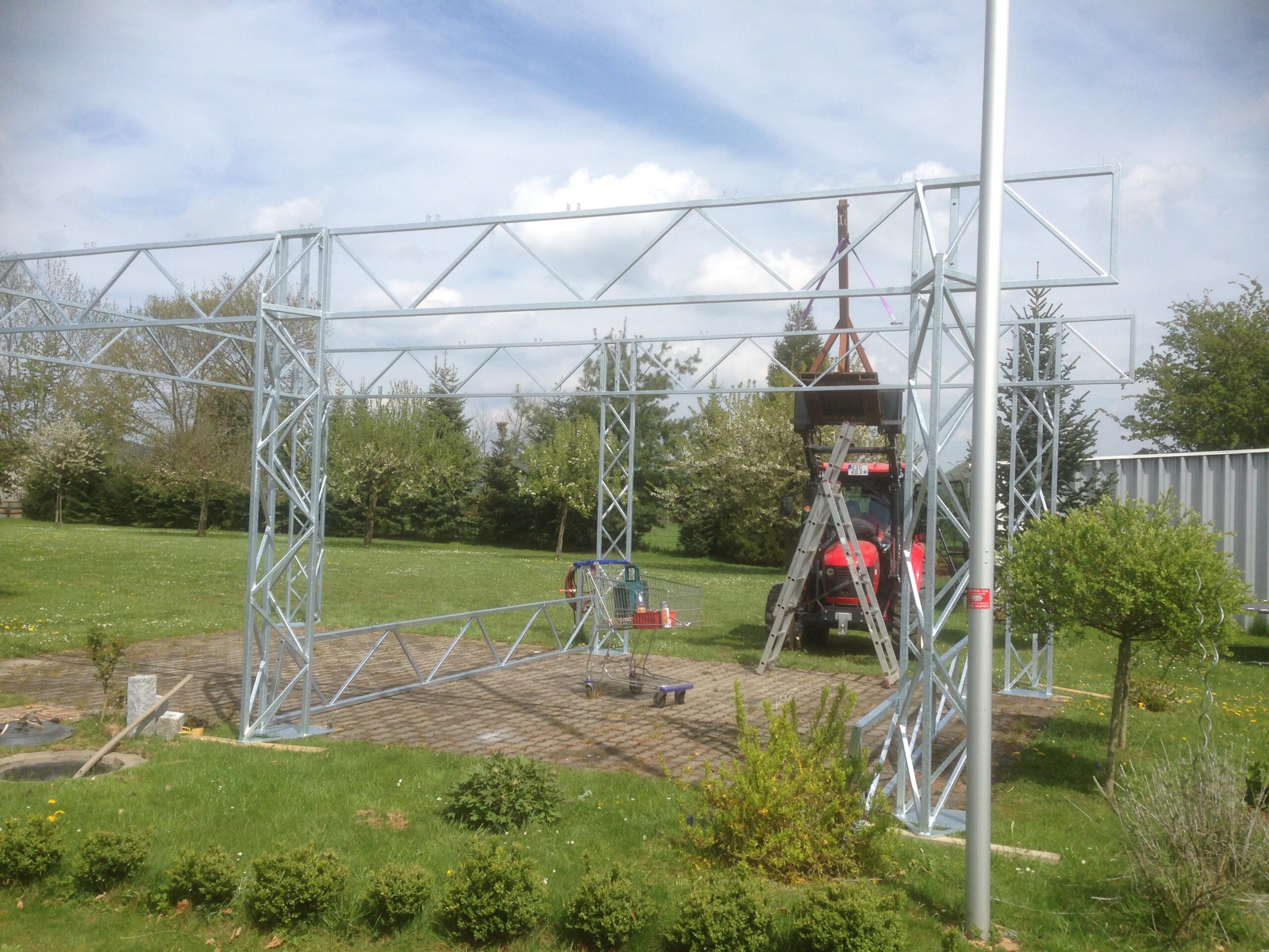 Teilmontierte verzinkte Stahlfachwerkkonstruktion von Stahlbau von Stahl- und Metallbau Drost auf einem gepflasterten Bereich, umgeben von Rasen und Bäumen, mit einem roten Traktor und einer Leiter im Hintergrund.
