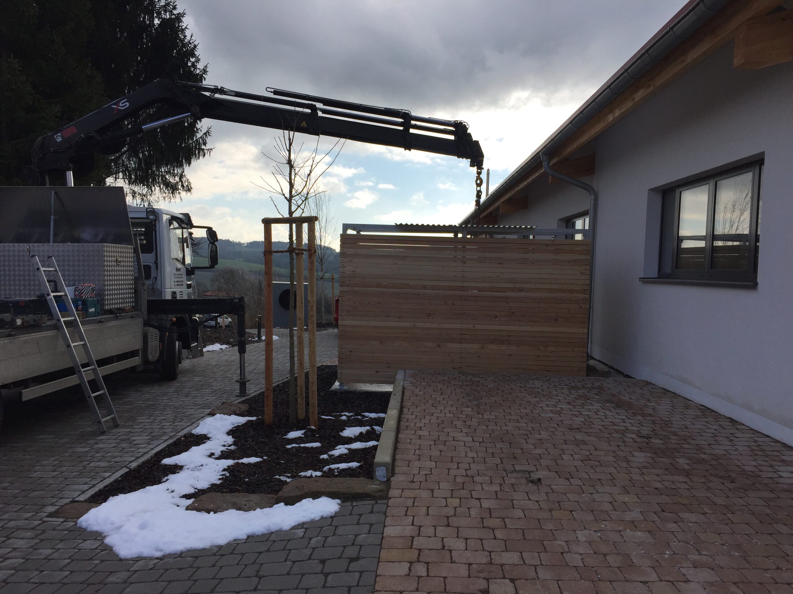 LKW mit Kran von Sonderanfertigungen von Stahl- und Metallbau Drost bei der Montage eines Holzsichtschutzes und der Gestaltung eines gepflasterten Außenbereichs neben einem modernen Haus.