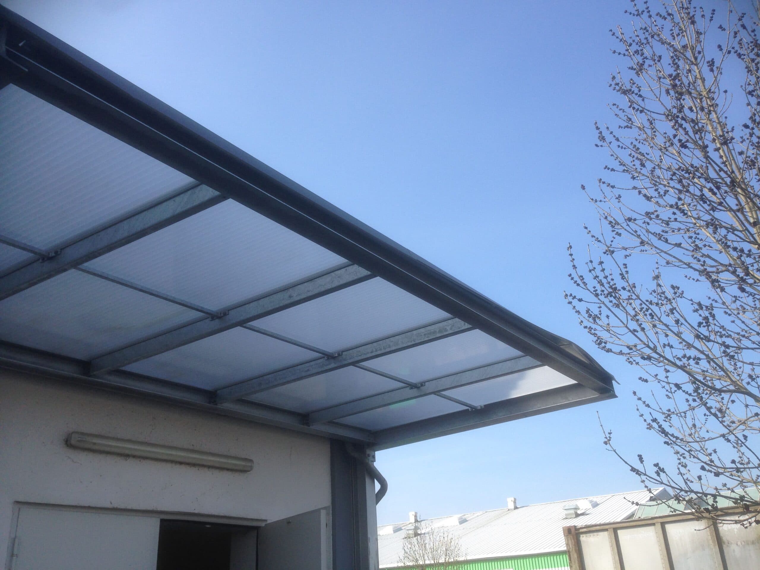 Ein Vordach von Stahl- und Metallbau Drost mit einer robusten Metallkonstruktion und einer transparenten Wellplattenabdeckung, montiert an einem Gebäude. Der blaue Himmel und ein kahler Baum sind im Hintergrund sichtbar.