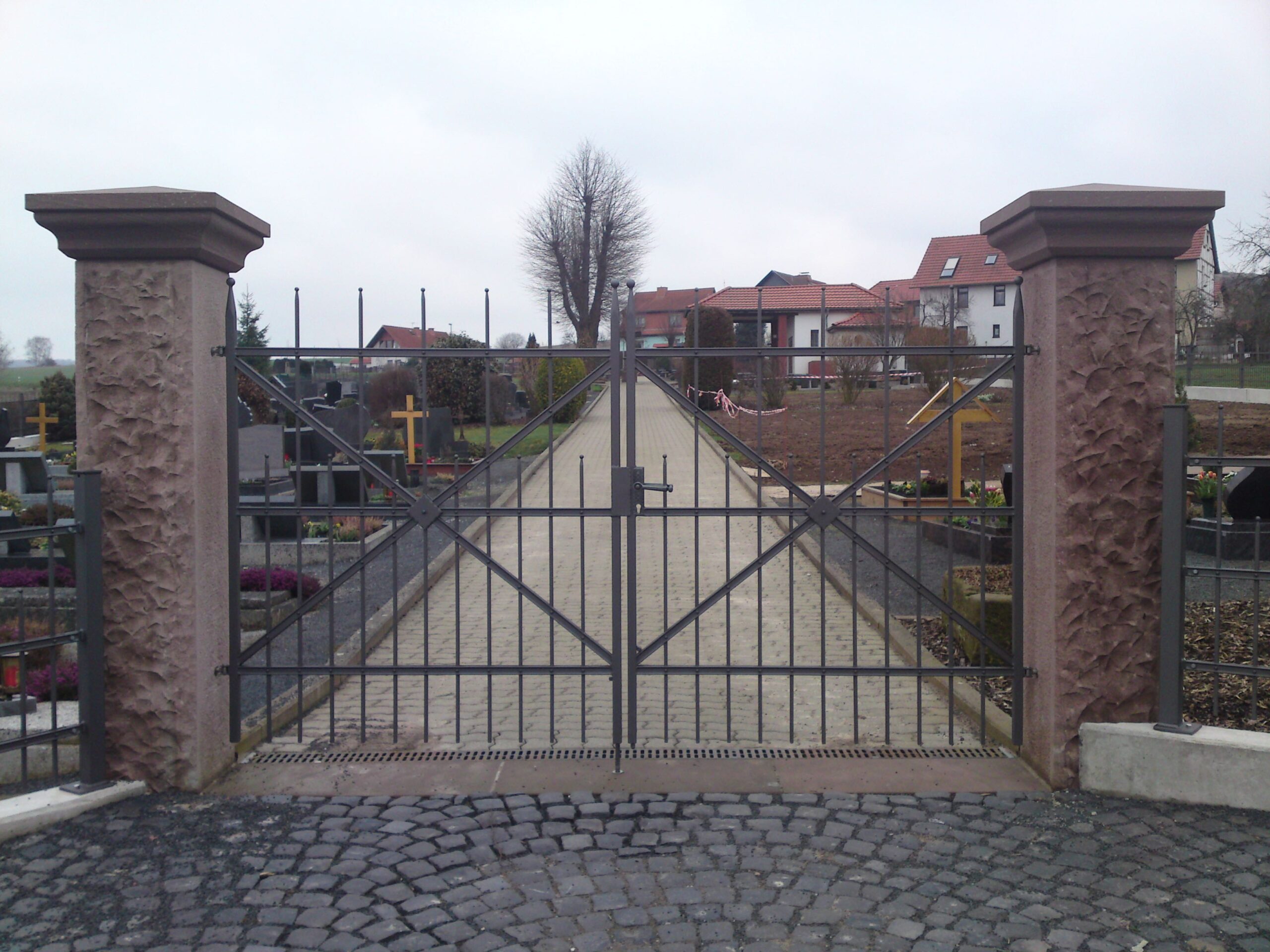 Ein geschlossenes, robustes, doppelflügeliges Metalltor in Dunkelgrau, mit vertikalen Gitterstäben und diagonalen Verstrebungen, flankiert von zwei massiven, strukturierten Steinpfeilern. Das Tor öffnet sich zu einem gepflasterten Weg, im Hintergrund sind Gräber und Gebäude sichtbar. Ein Qualitätsprodukt von Tore & Torsysteme von Stahl- und Metallbau Drost.