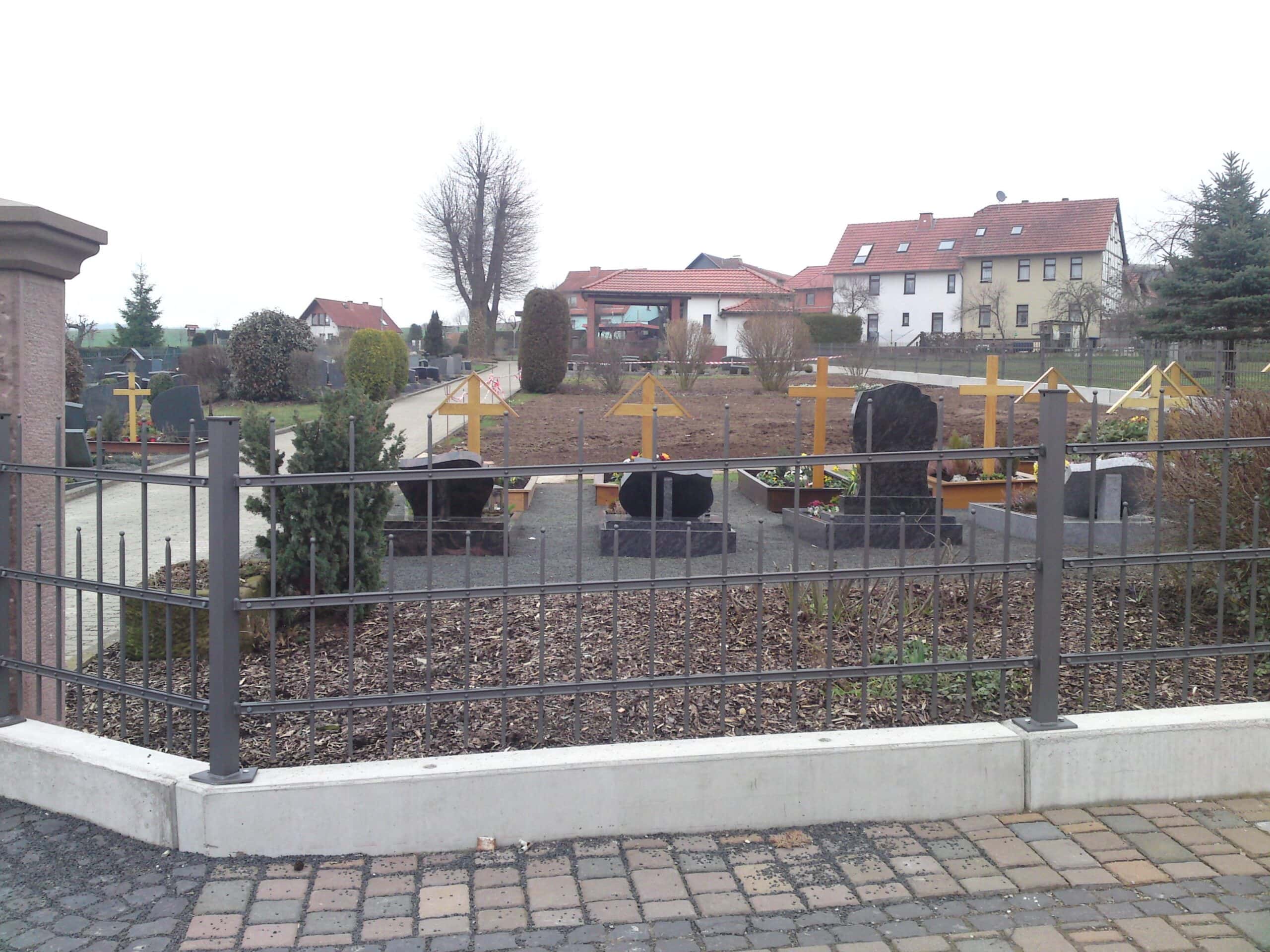Dunkelgrauer Metallzaun von Zaunbau von Stahl- und Metallbau Drost umgibt einen Friedhof mit Gräbern und Holzkreuzen. Der Zaun steht auf einem Betonsockel, im Hintergrund sind Häuser zu sehen.