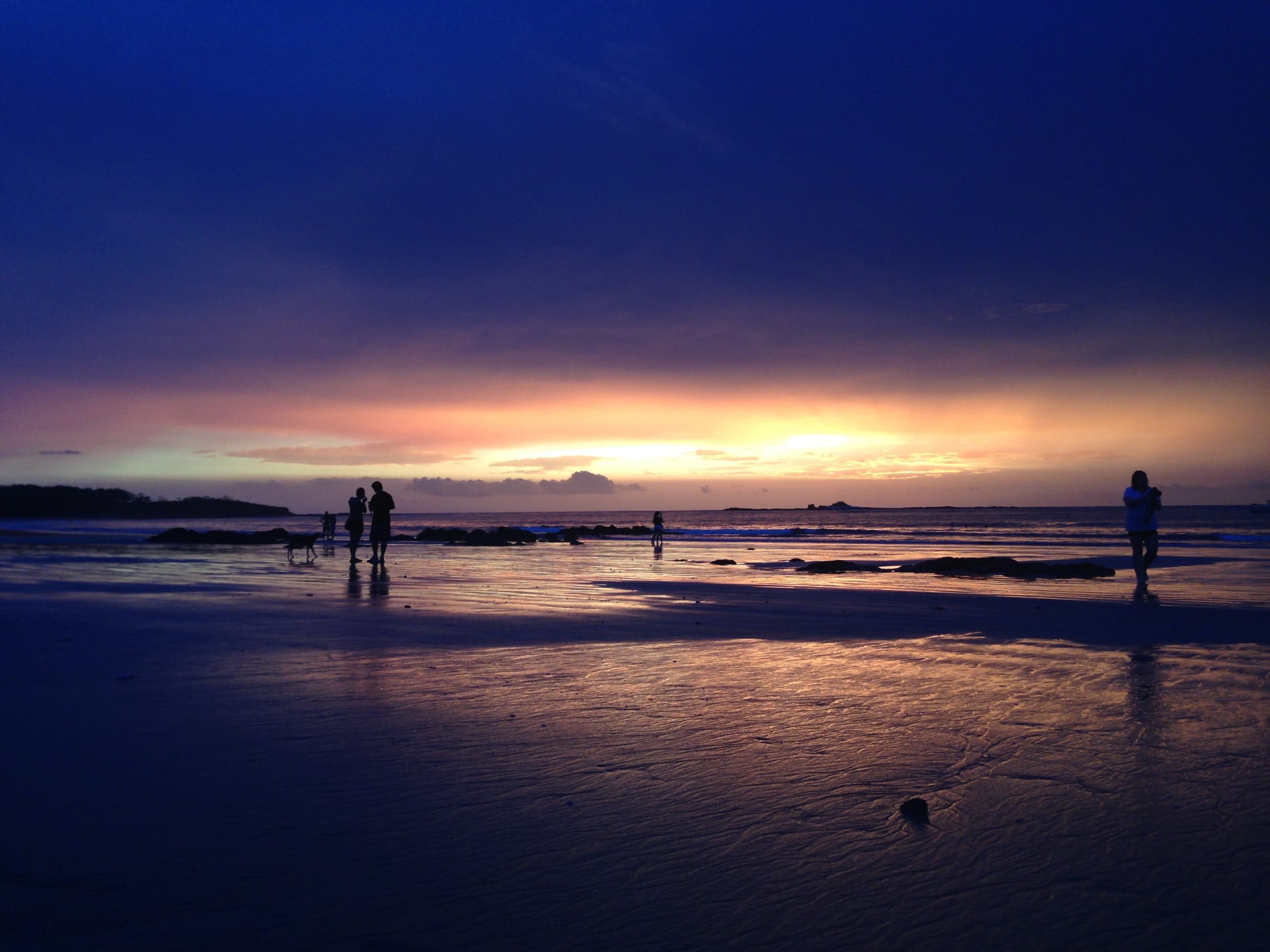 A dark blue sunset on the beach, with only a strip of sunlight visible at the horizon line in Tamarindo, Costa Rica. Two shadows figures are visible on the beach.