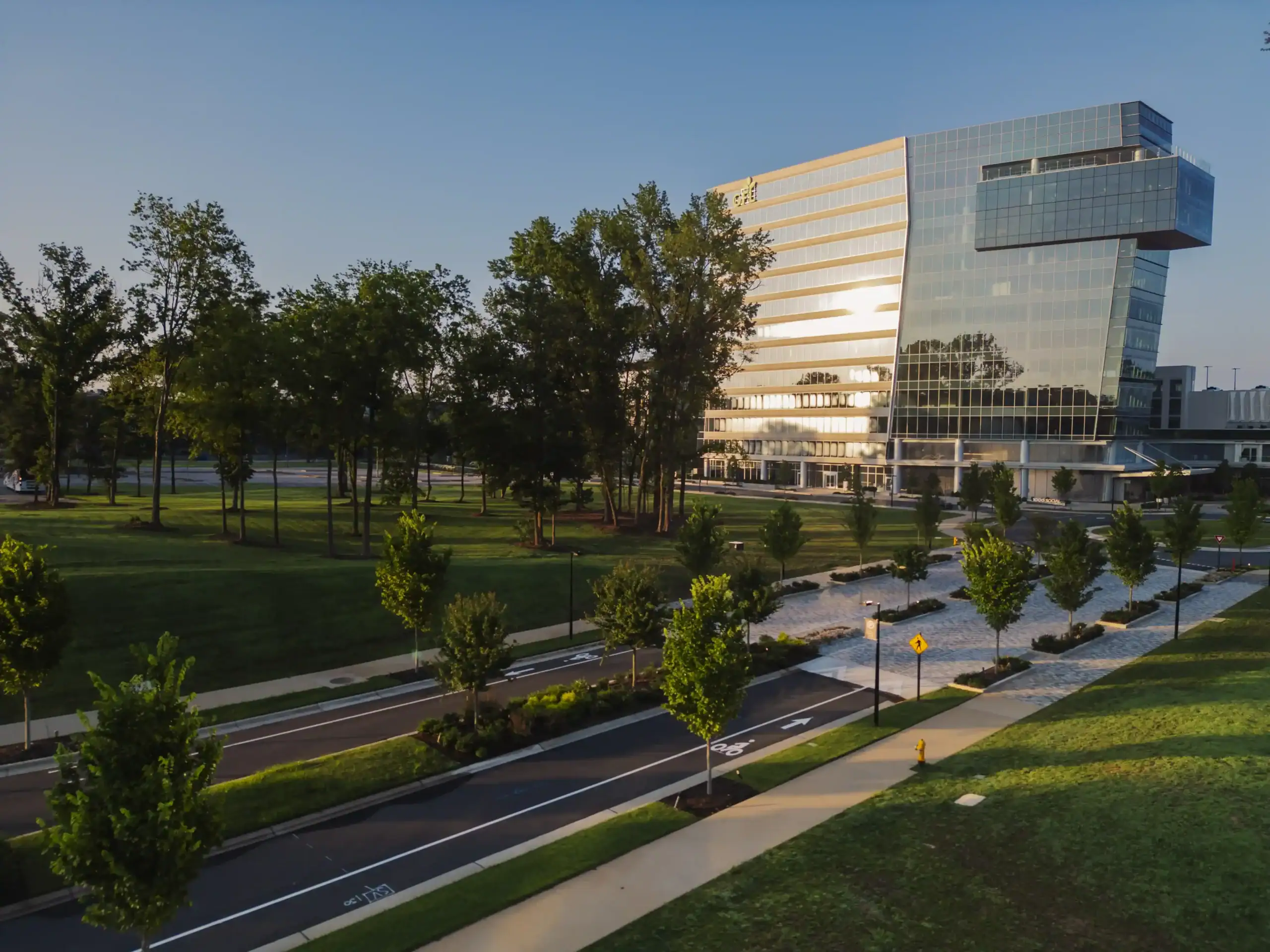 Modern glass Raleigh office building surrounded by landscaped green space, tree-lined streets, pedestrian walkways, and bike lanes during golden hour.