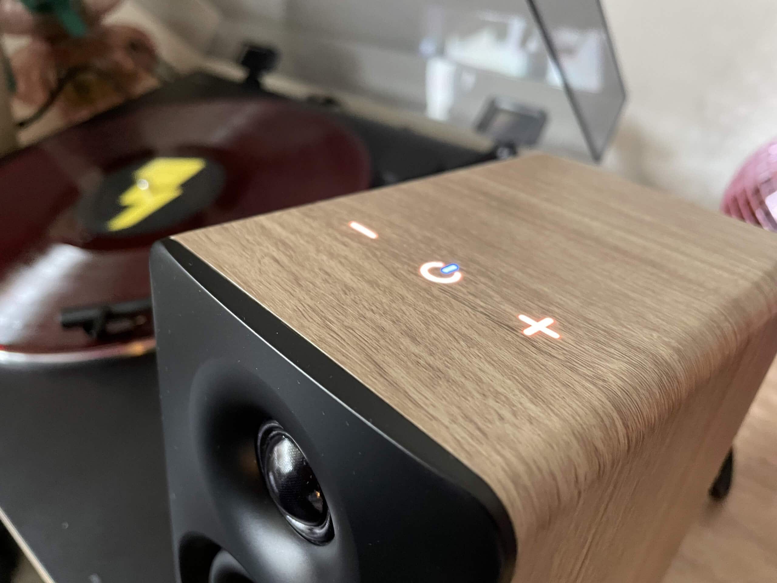 Picture showing the control panel, on the top face of the right-hand speaker, lit up. A minus symbol, a power symbol lit with a blue segment to indicate a Bluetooth connection, and a plus symbol.