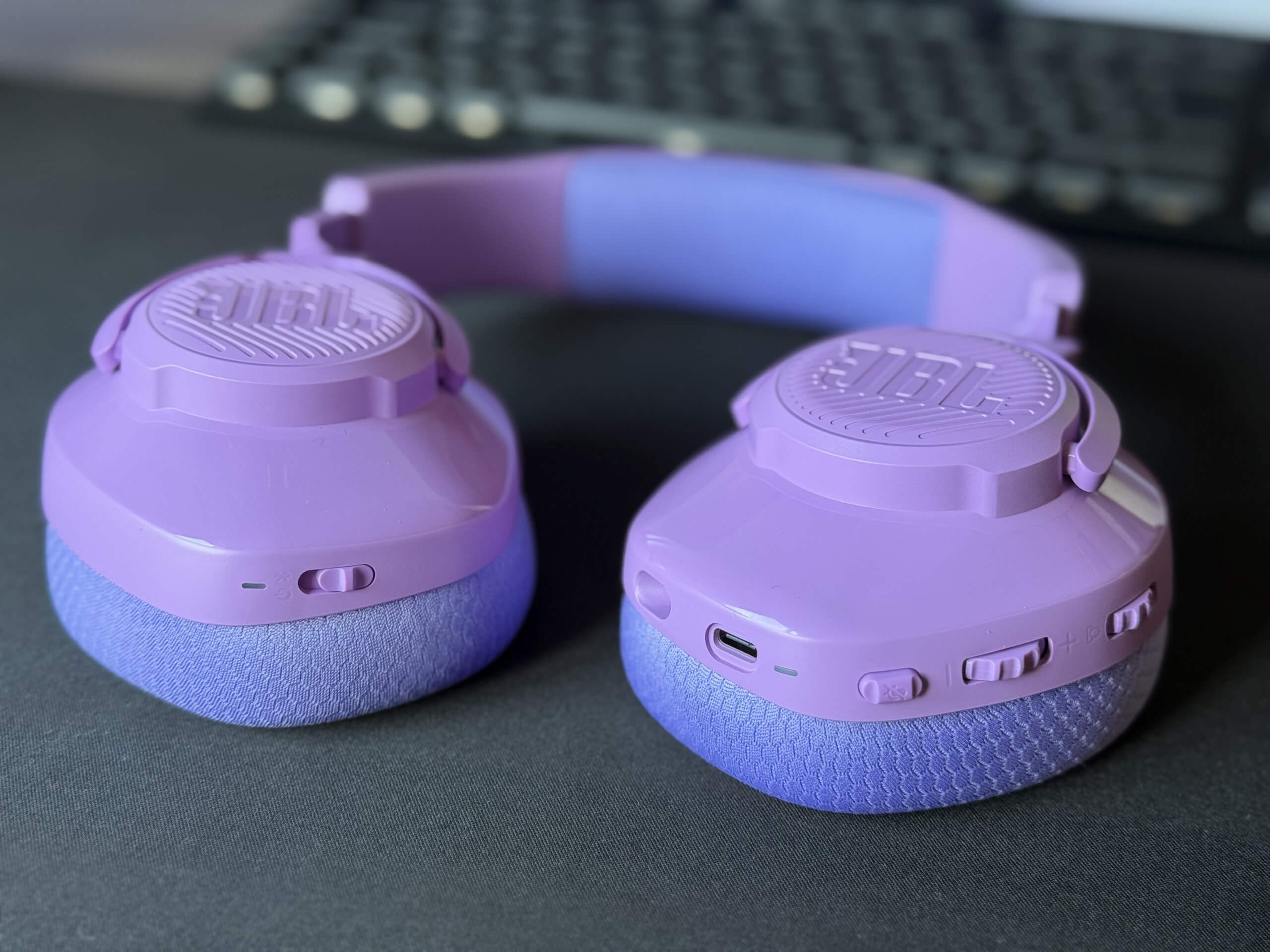 Purple JBL Quantum 360 wireless headphones laying flat on a desk with a black mouse mat below and a blurred keyboard in the background.